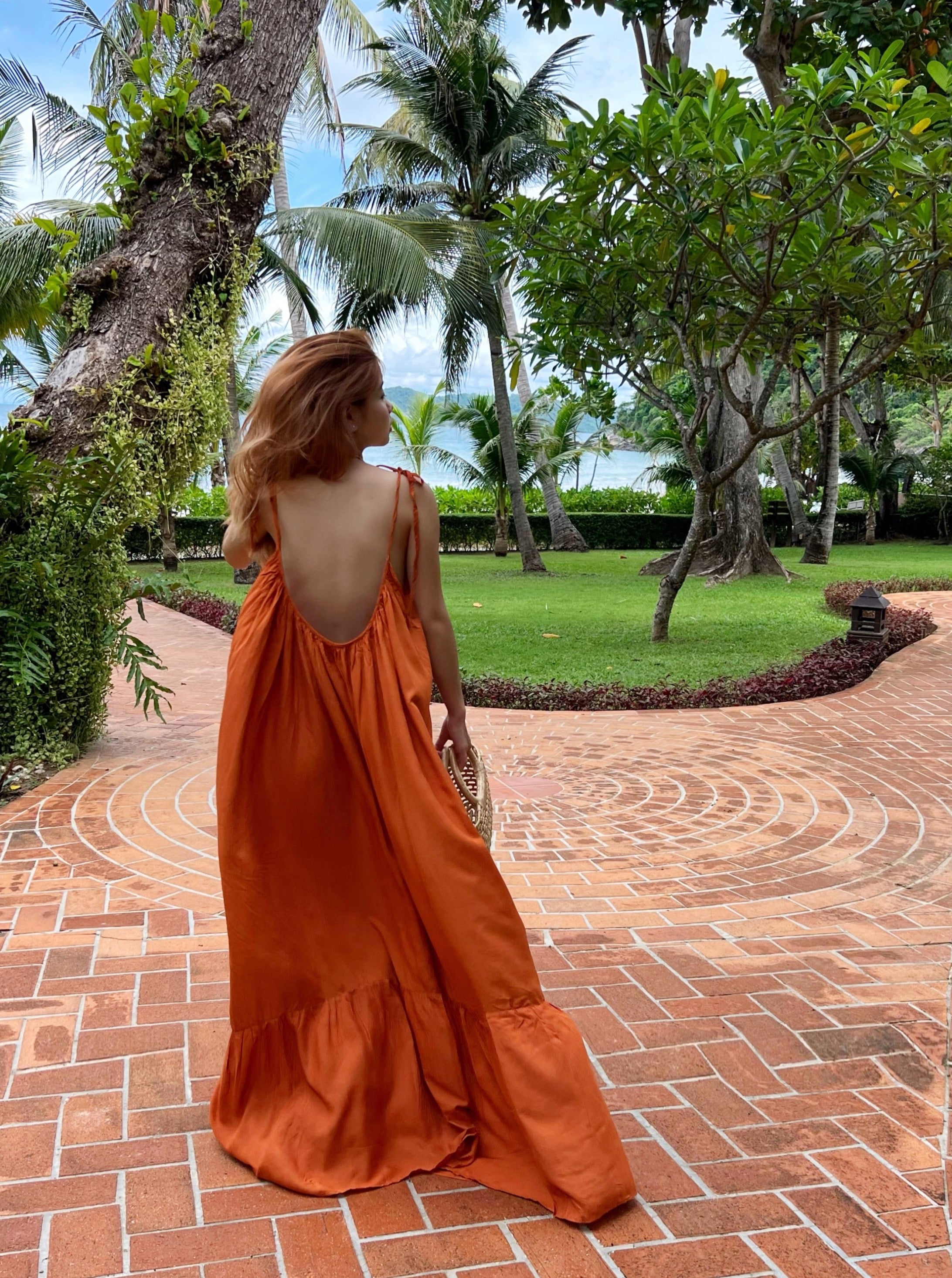 A person wearing a rust backless maxi dress with tie-shoulder design and ruffle hem detailing, standing on a pathway in a tropical resort setting.