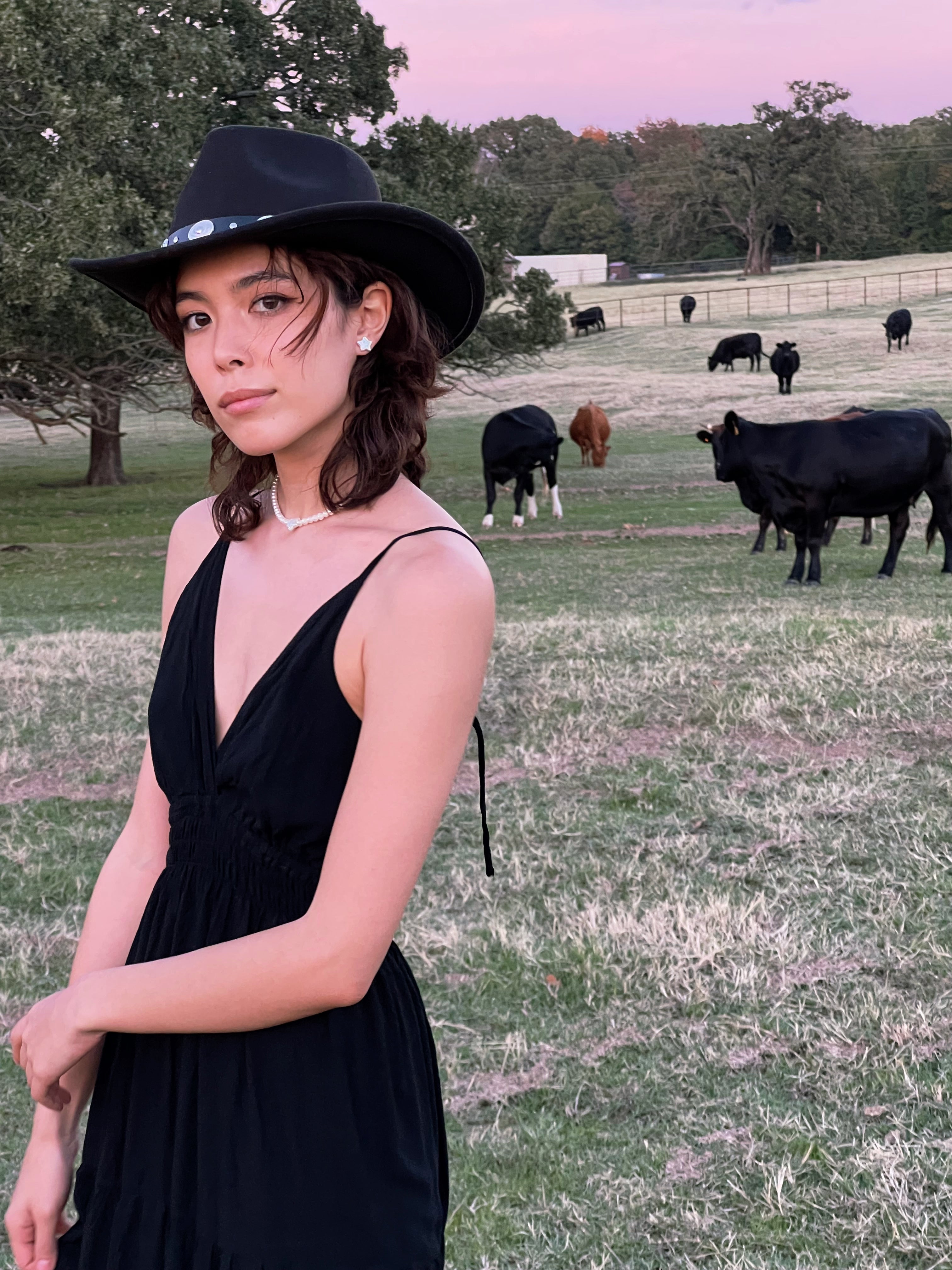 A woman standing in a field with a cow in the background, wearing a black maxi dress with a smocked bodice.