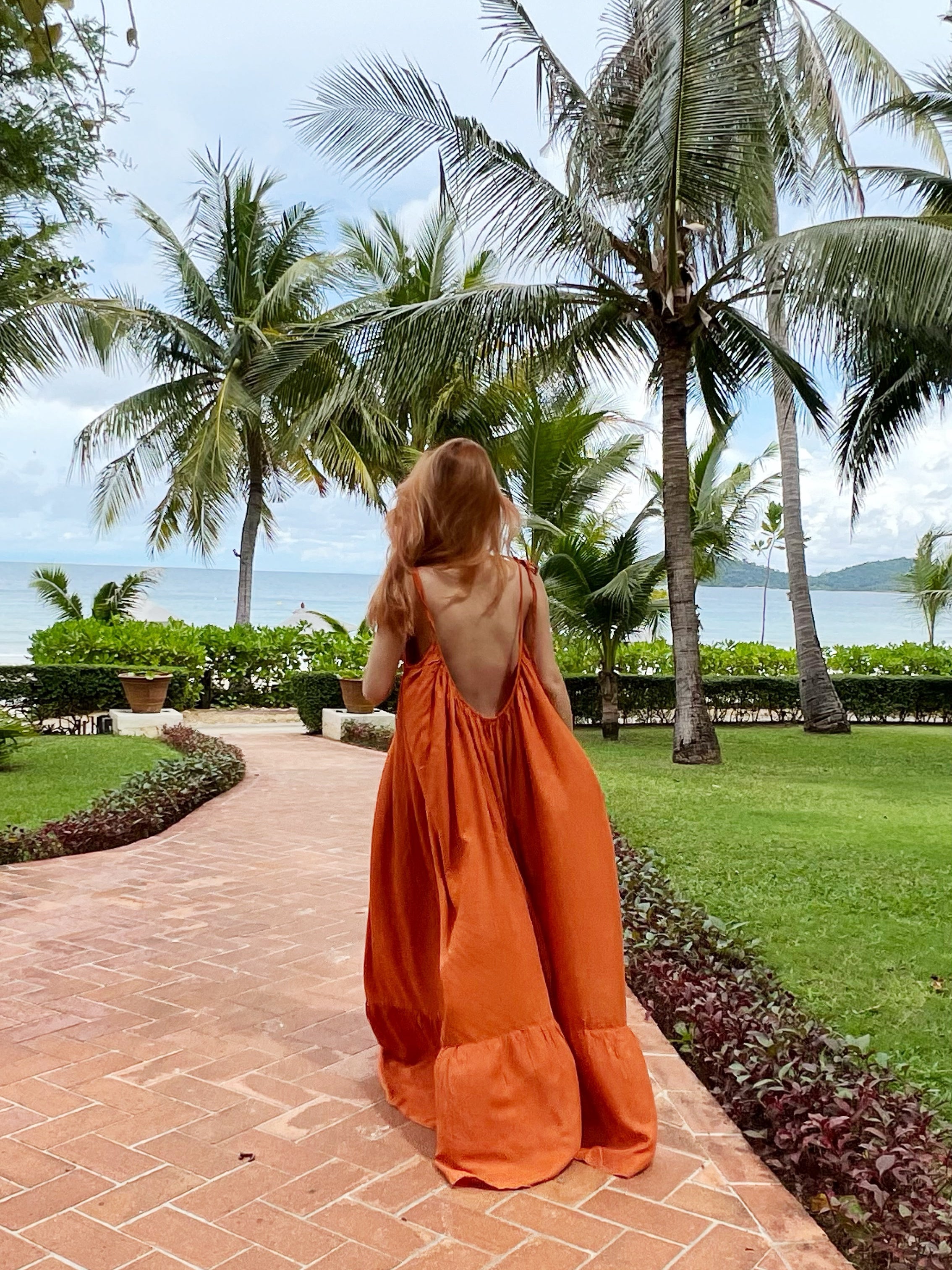 A person wearing a rust backless maxi dress with tie-shoulder design and ruffle hem detailing, standing on a pathway in a tropical resort setting.