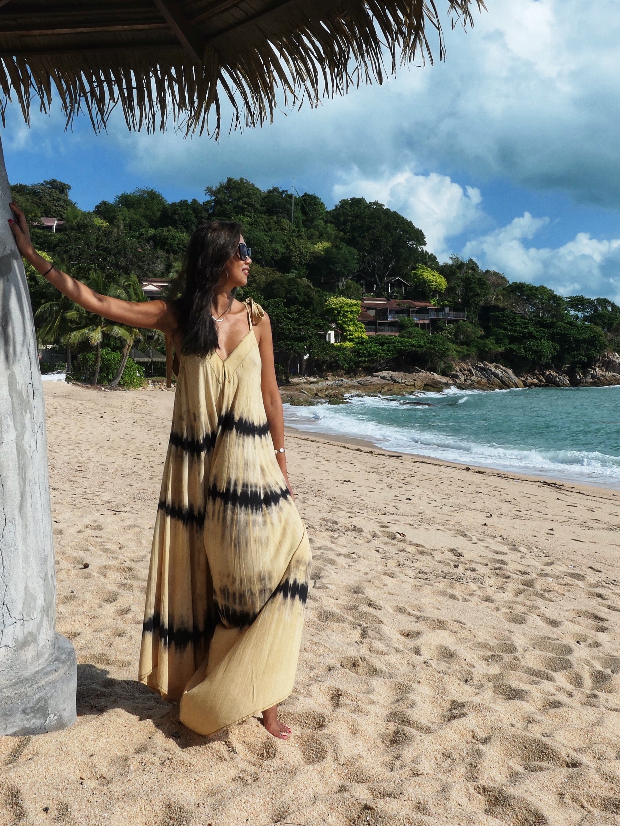 Woman in tie dye jumpsuit with an oversized fit and back-V silhouette, perfect for yoga, beach cover-up, or casual day-to-night wear by Coco De Chom standing on a sandy beach with ocean and rocks in the background.