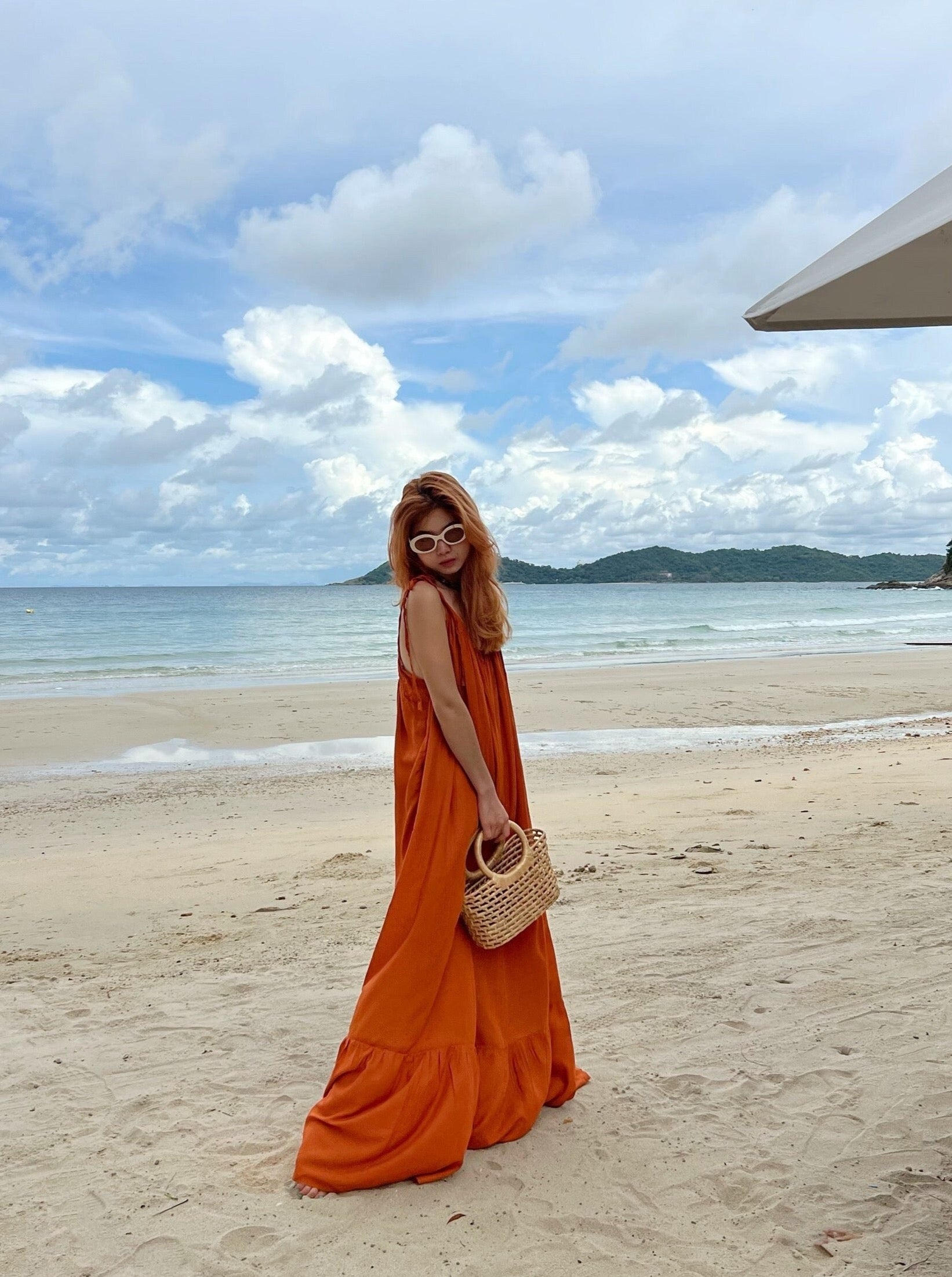 A person wearing a rust backless maxi dress with tie-shoulder design and ruffle hem detailing, standing on a pathway in a tropical resort setting.