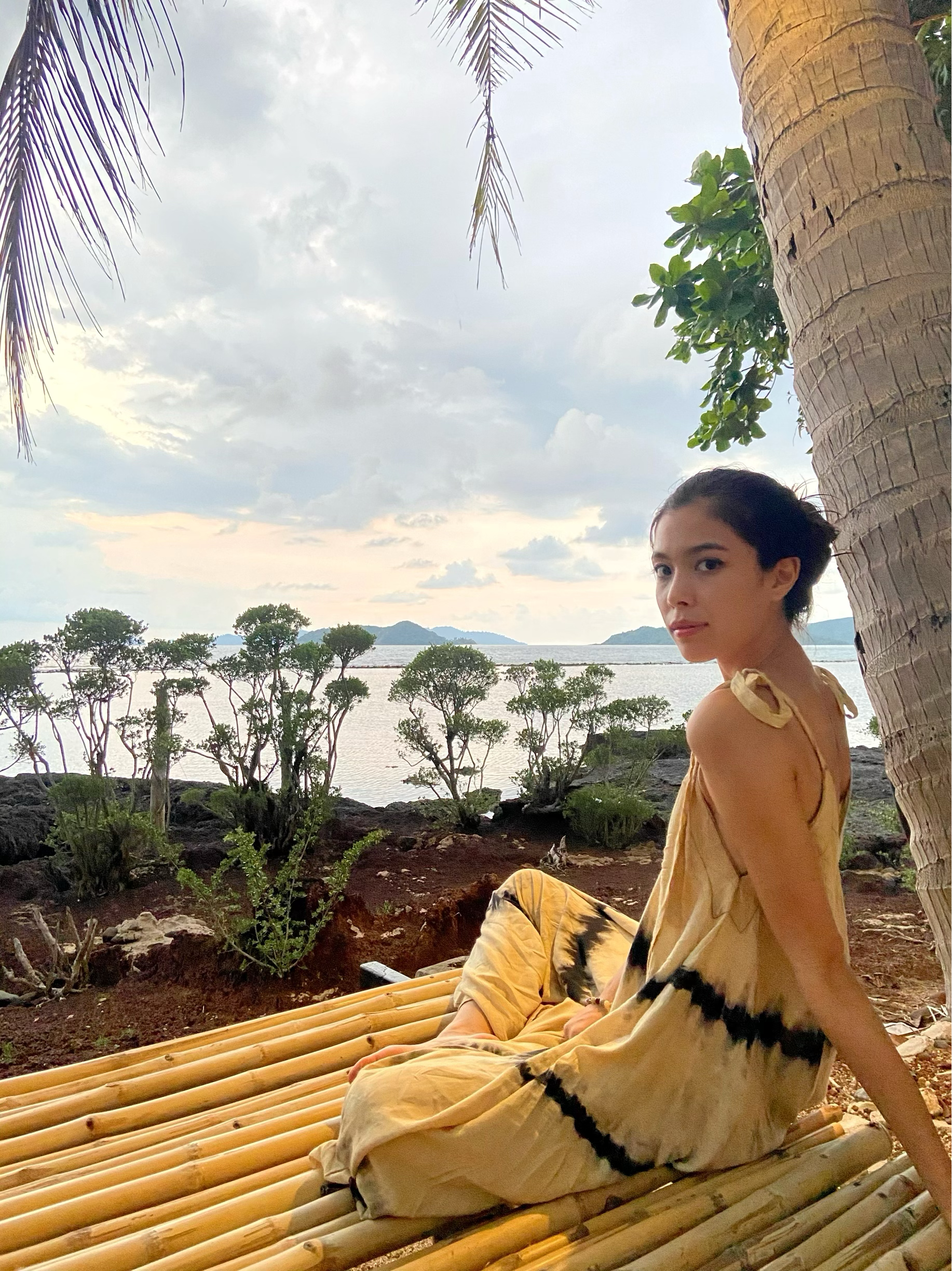 Woman in tie dye jumpsuit with an oversized fit and back-V silhouette, perfect for yoga, beach cover-up, or casual day-to-night wear by Coco De Chom standing on a sandy beach with ocean and rocks in the background.