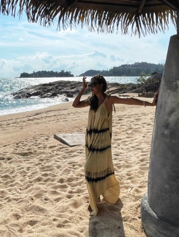 Woman in tie dye jumpsuit with an oversized fit and back-V silhouette, perfect for yoga, beach cover-up, or casual day-to-night wear by Coco De Chom standing on a sandy beach with ocean and rocks in the background.