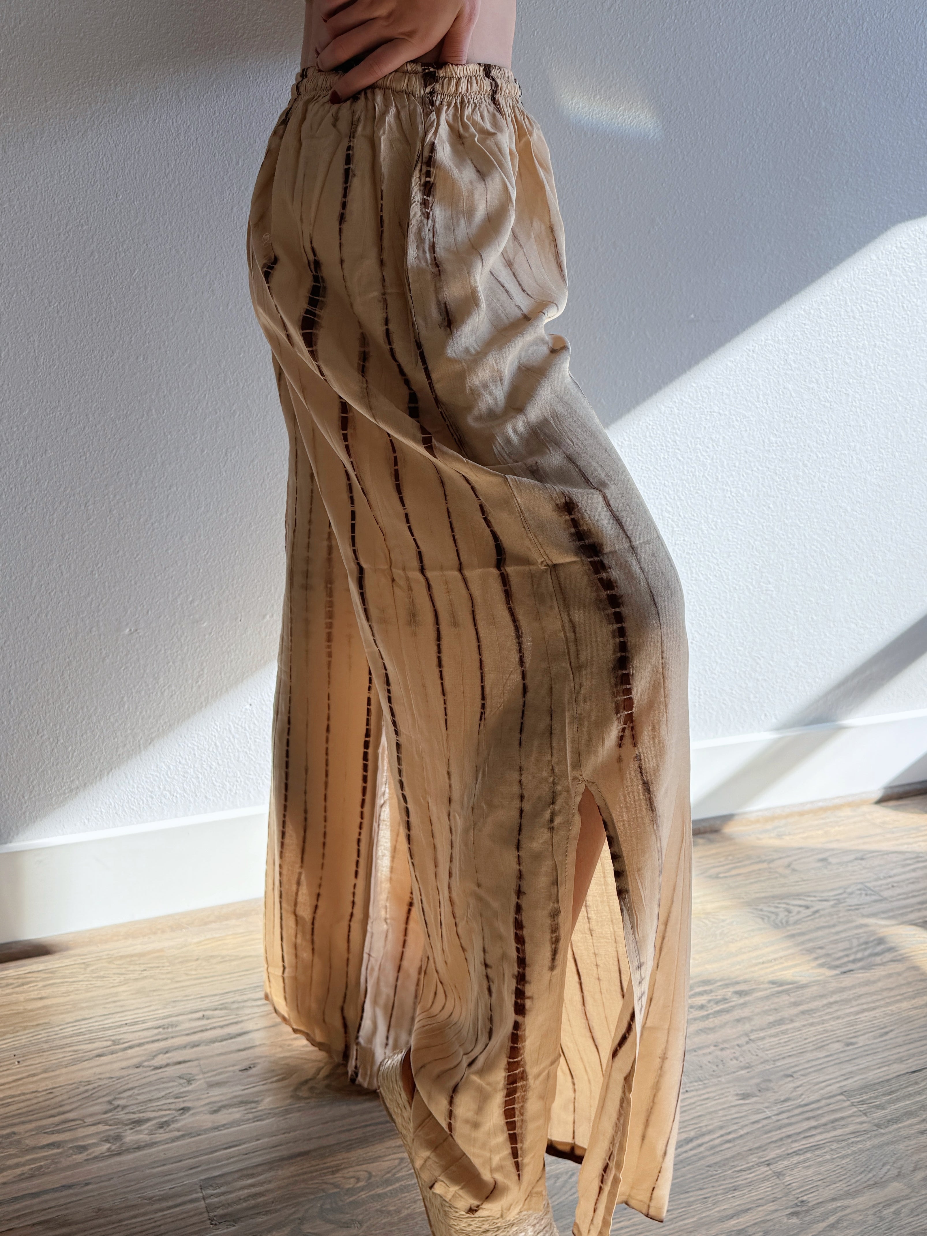 Woman wearing a white top and beige tie dye wide-leg pants, giving a look boho vibe perfect outfit for beach vacation, standing in a room with sunlight streaming in.