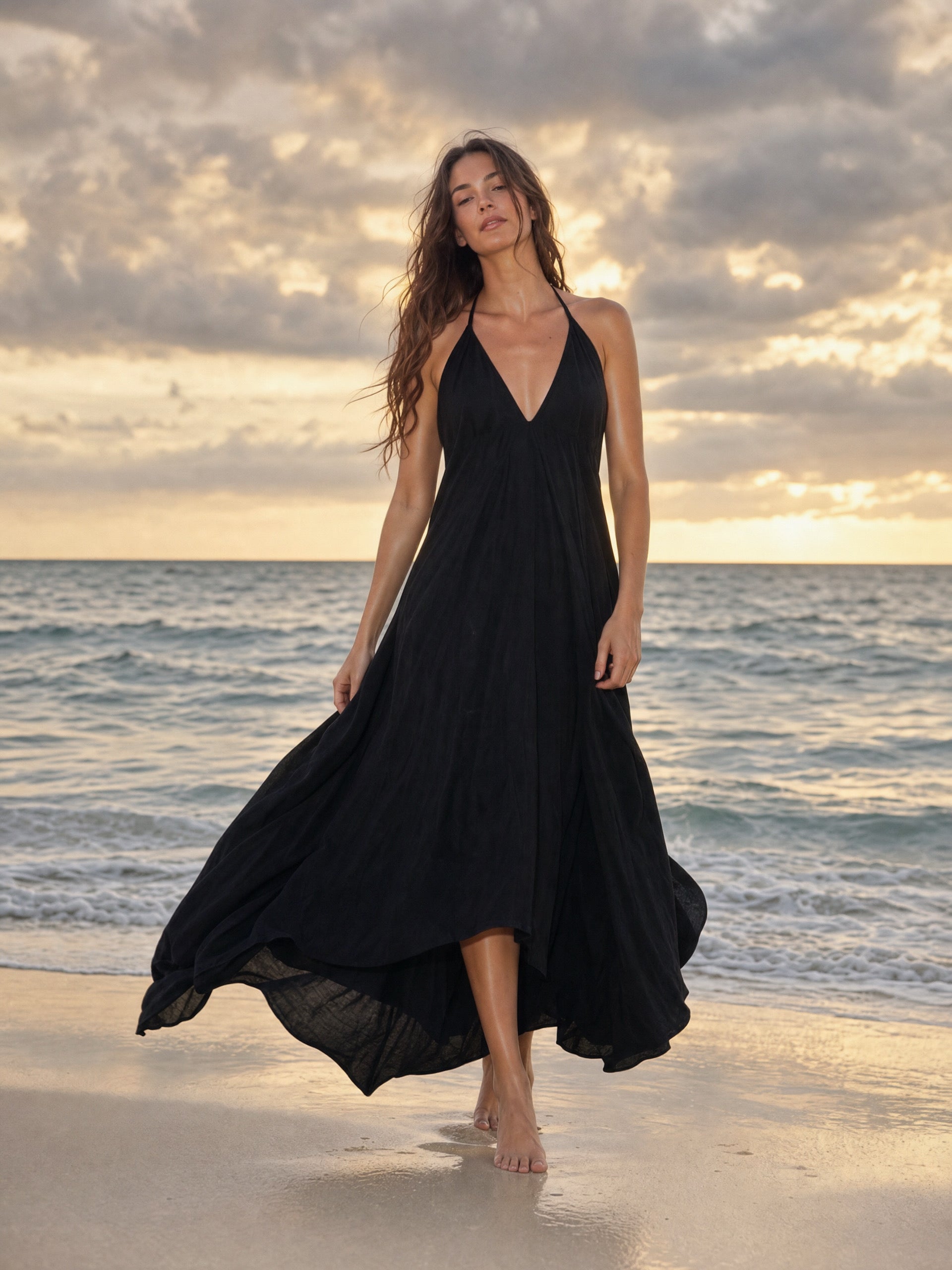 Woman walking on the beach at dusk wearing the Flamingo Maxi Dress in solid black, plunging V-neckline and high-low hem