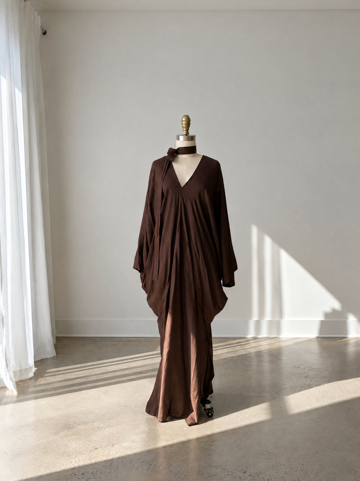 A dark brown draped maxi dress displayed on a mannequin in a minimalist studio setting with natural light casting shadows on the wall and floor.