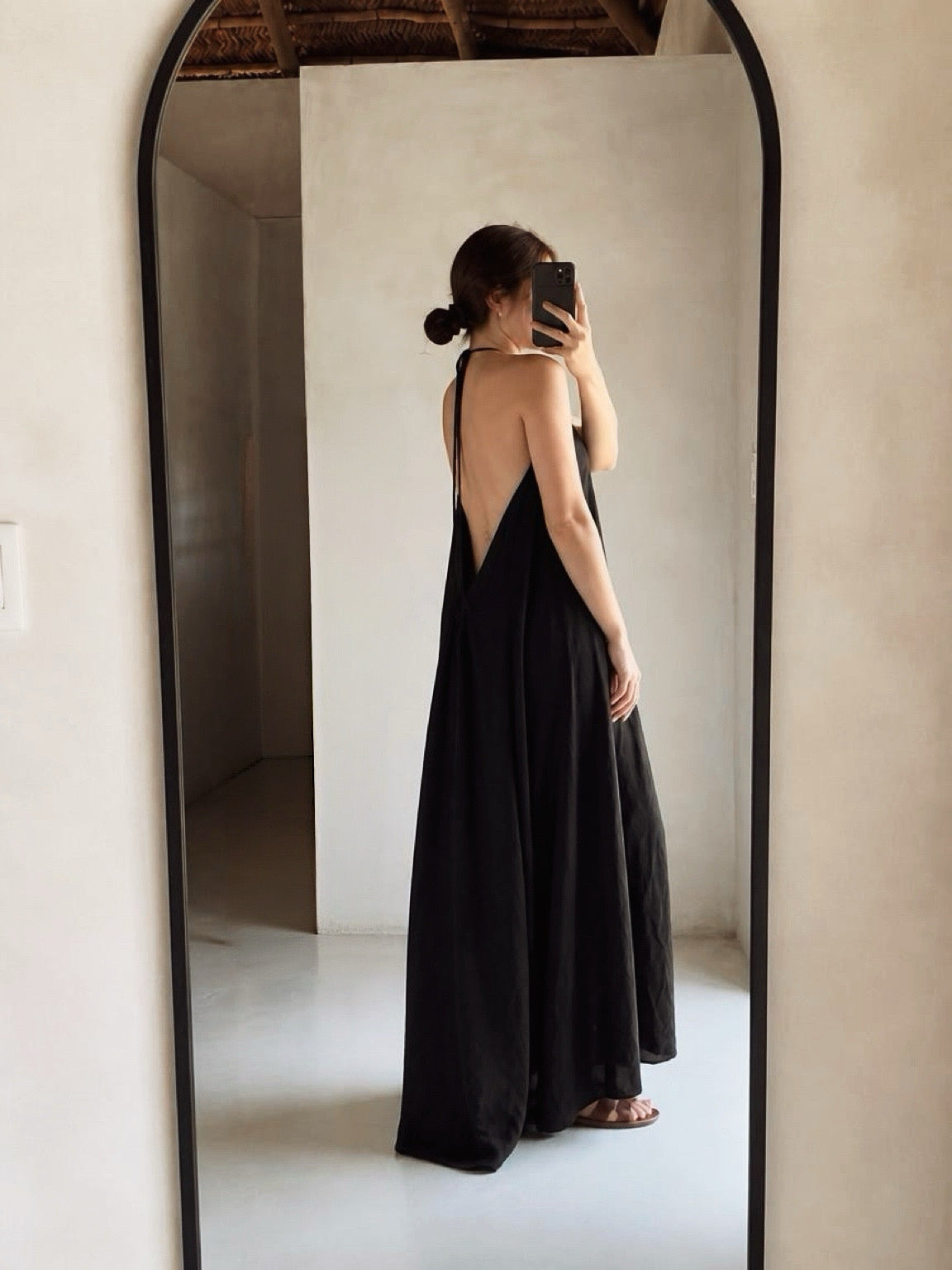 A woman takes a mirror selfie showing the back of a long black maxi dress with a deep V-back and halter neckline, set against a minimalist beige interior.