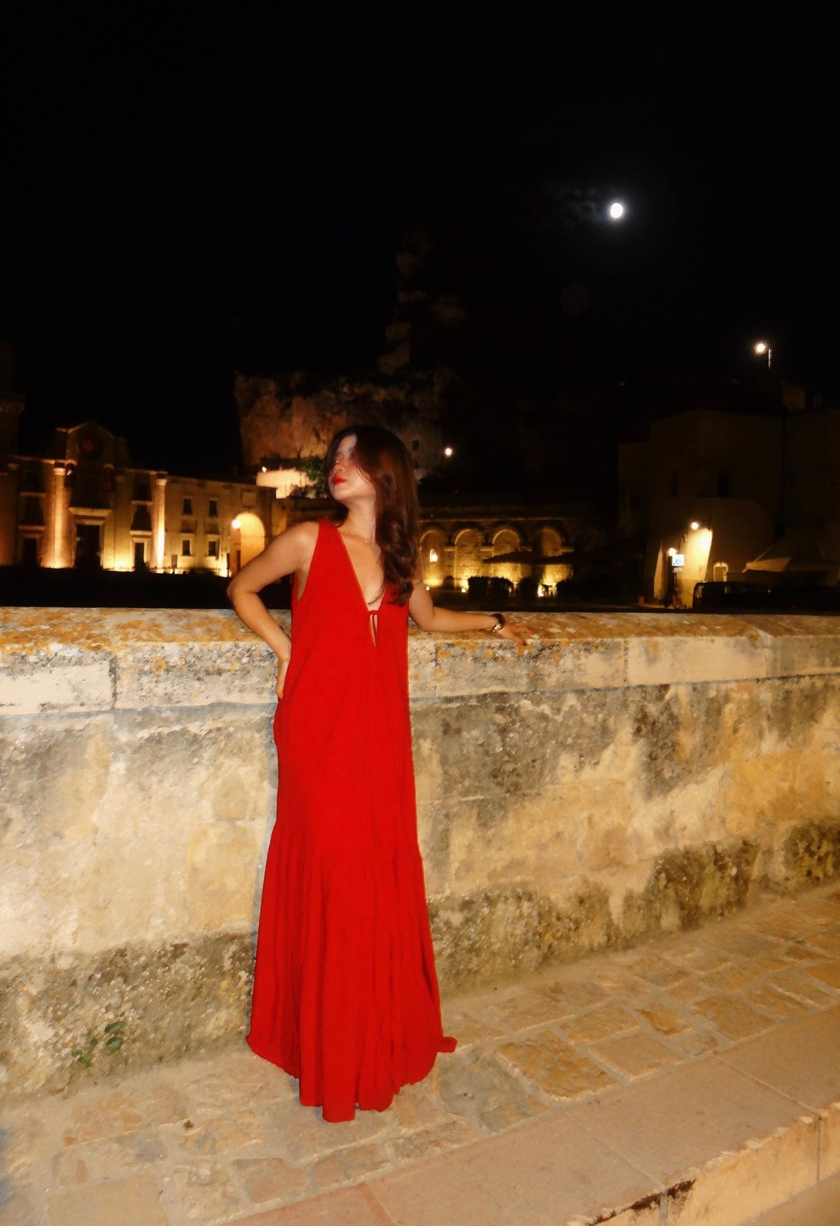Woman in a red maxi dress standing against a stone wall at night with moonlit sky.