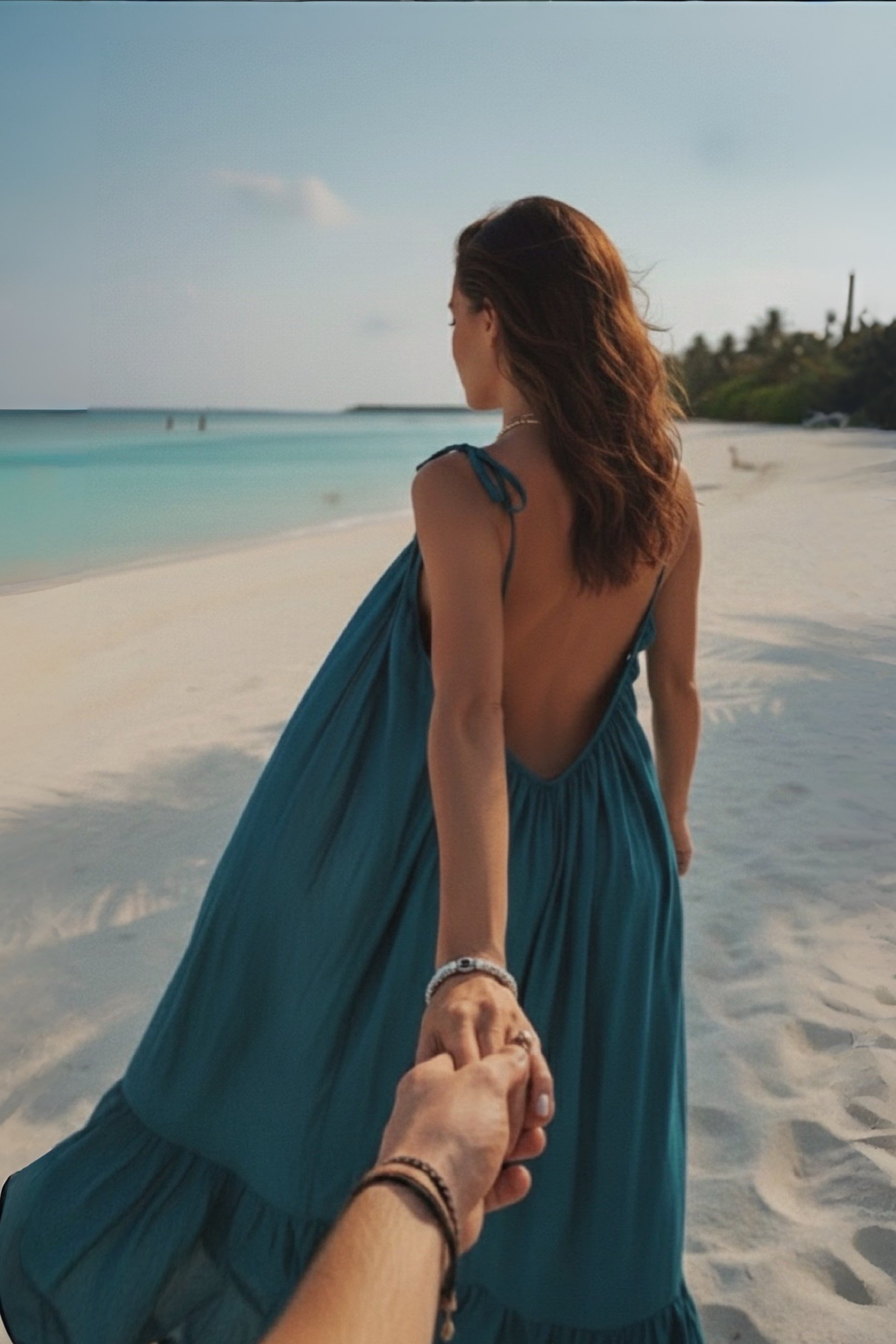 Woman in a teal open back maxi dress holding hands with another person on a beach.