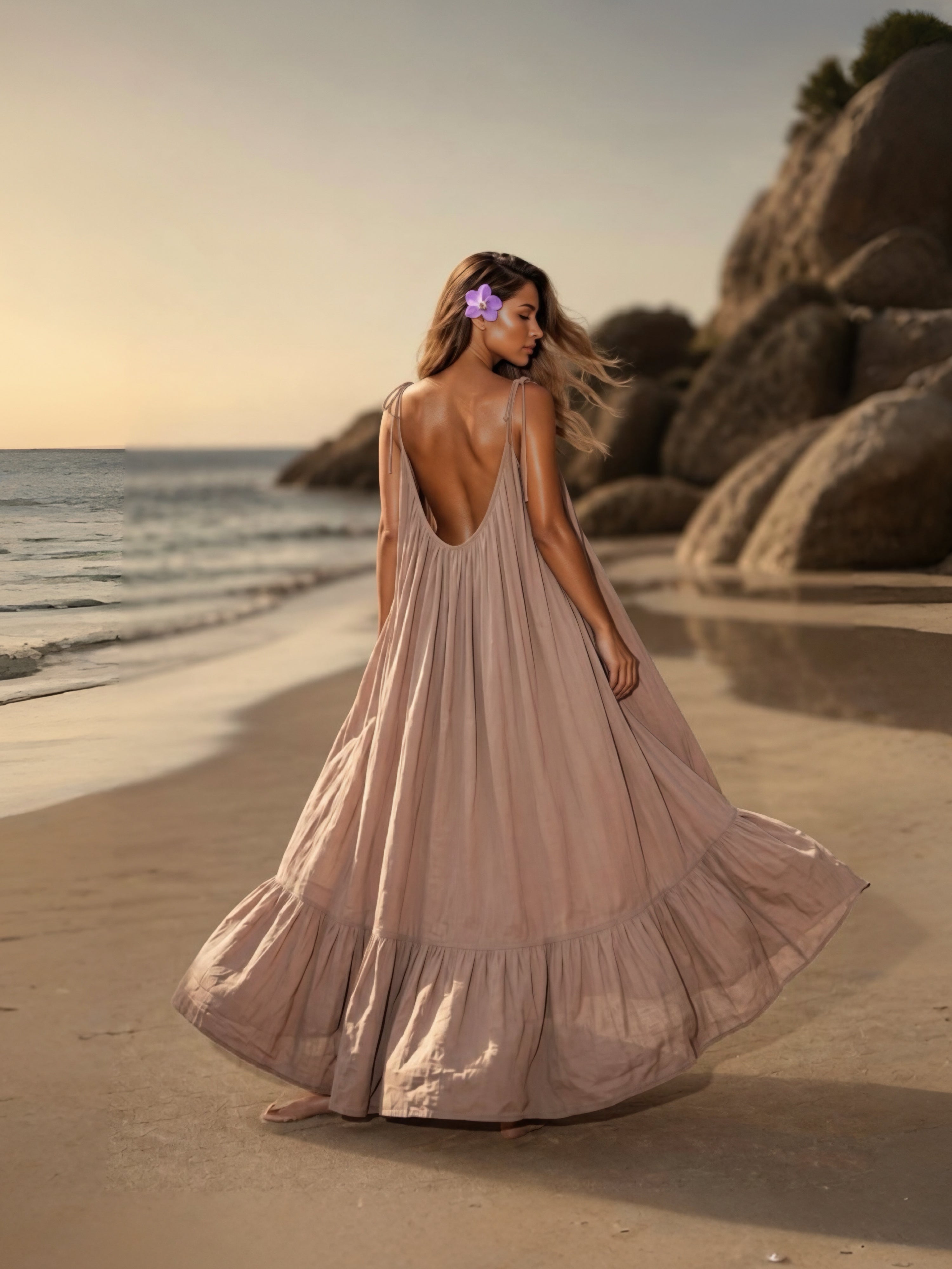 Woman in a flowing open back maxi dress in nude standing on a beach with large rocks and sunset in the background