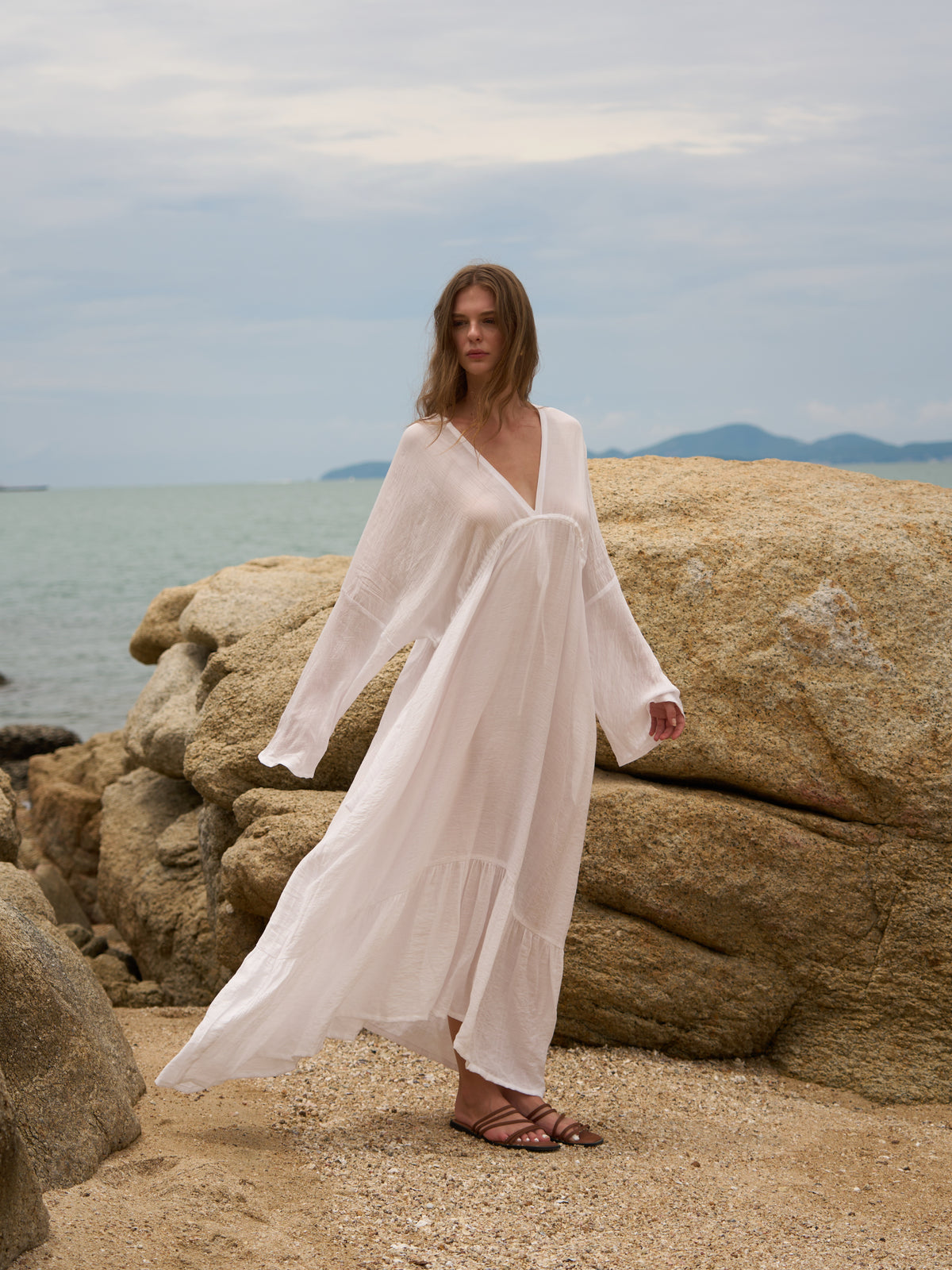 A woman wearing a flowing white long-sleeve maxi cover-up dress stands on a rocky beach with the sea and cloudy sky in the background.
