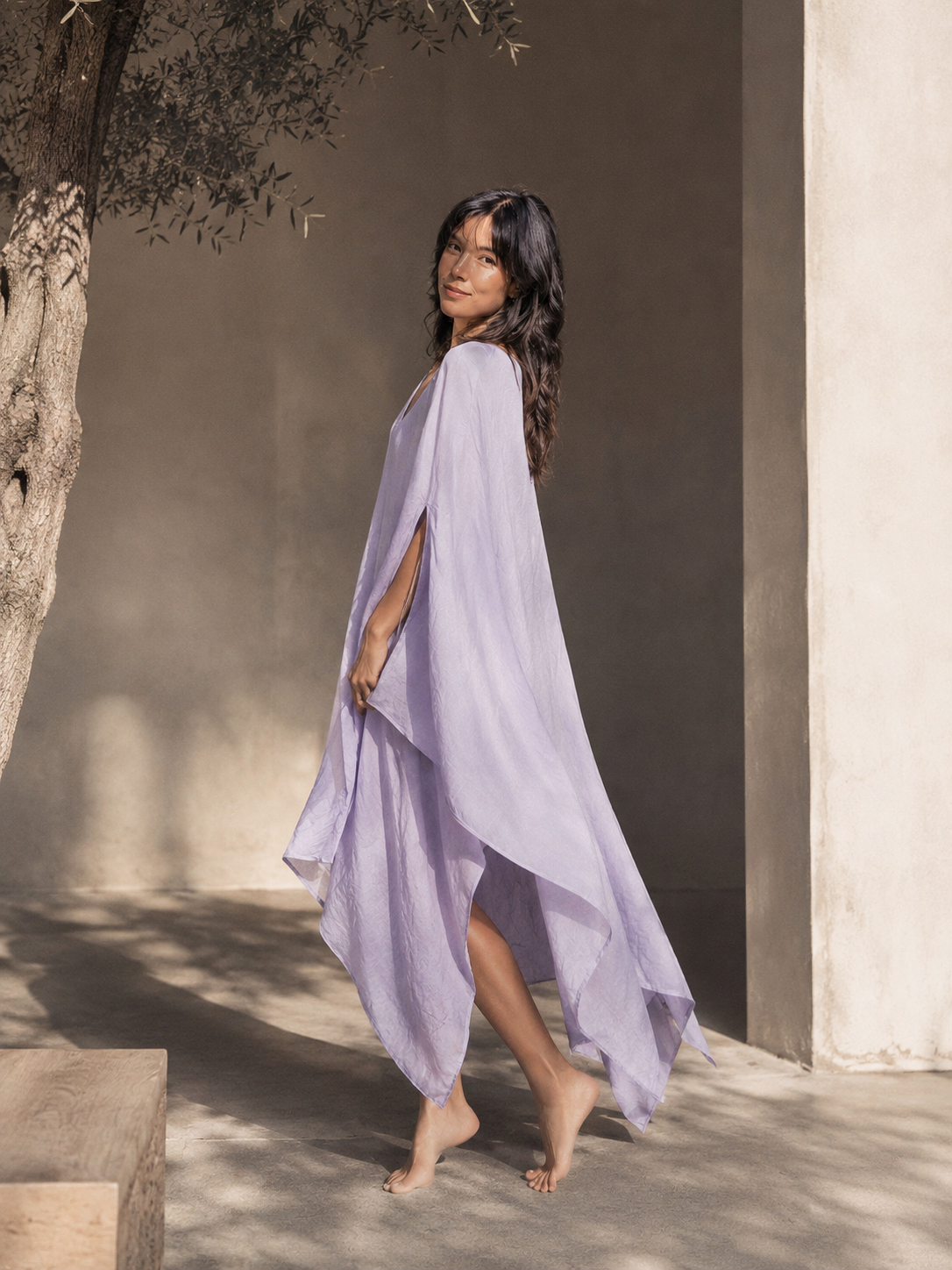 A woman wearing a flowing lavender high-short kaftan dress walks barefoot in a sunlit courtyard with textured stucco walls and a tree trunk  for poolside lounging and cruise outfits