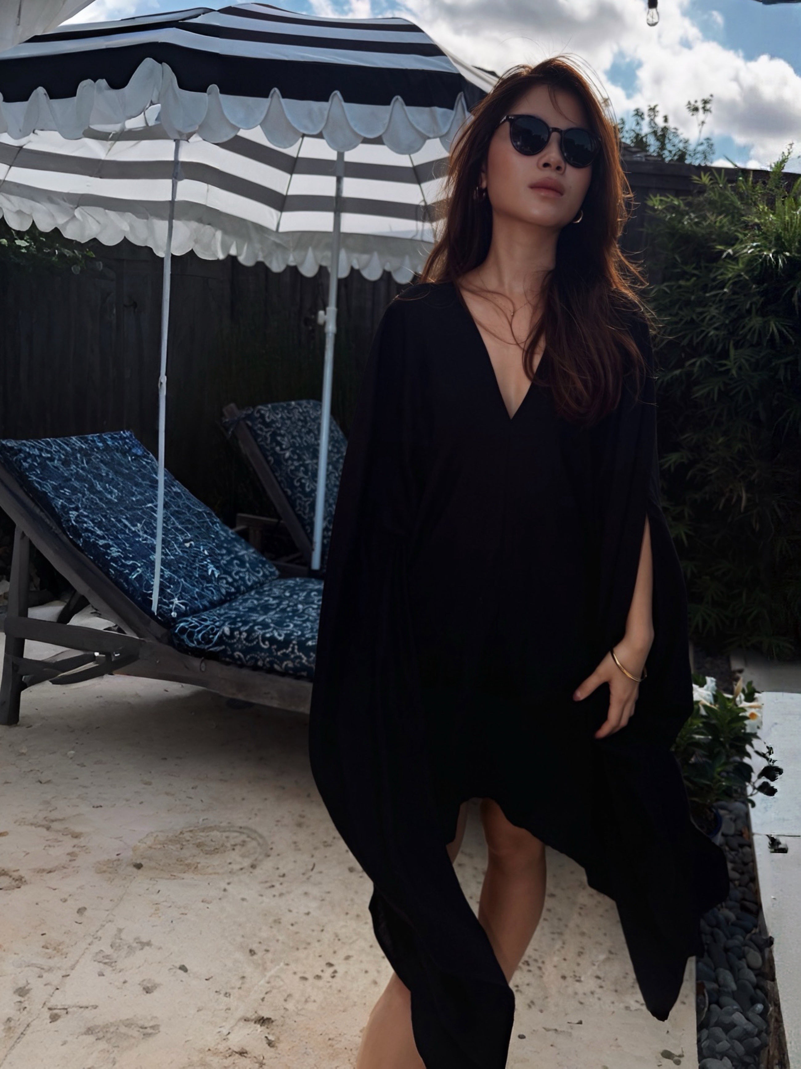 A woman wearing a black V-neck long-sleeve cover-up dress and sunglasses stands outdoors next to a black and white striped beach umbrella and lounge chairs.