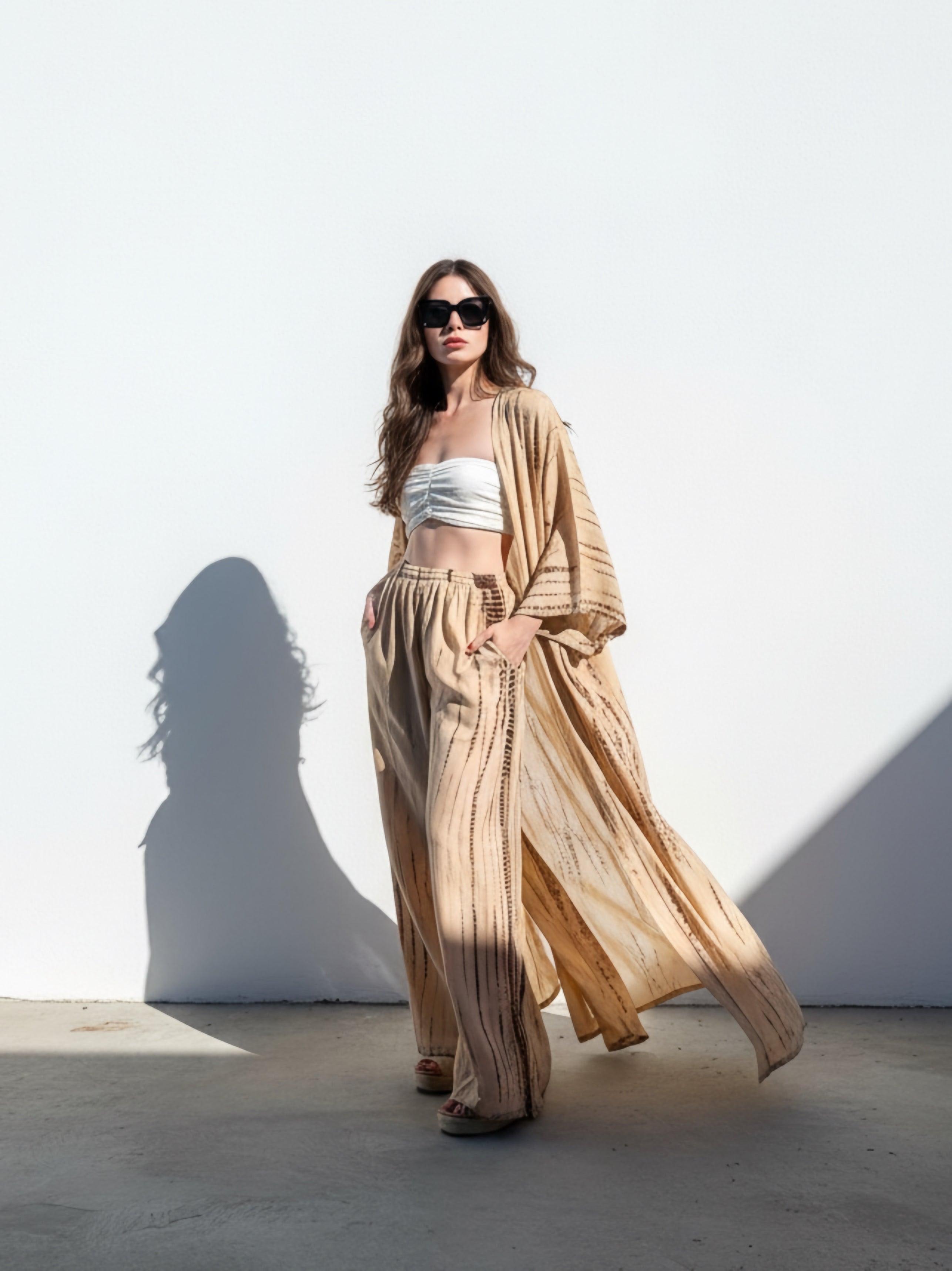 Woman wearing a white top and beige tie dye wide-leg pants, giving a look boho vibe perfect outfit for beach vacation, standing in a room with sunlight streaming in.