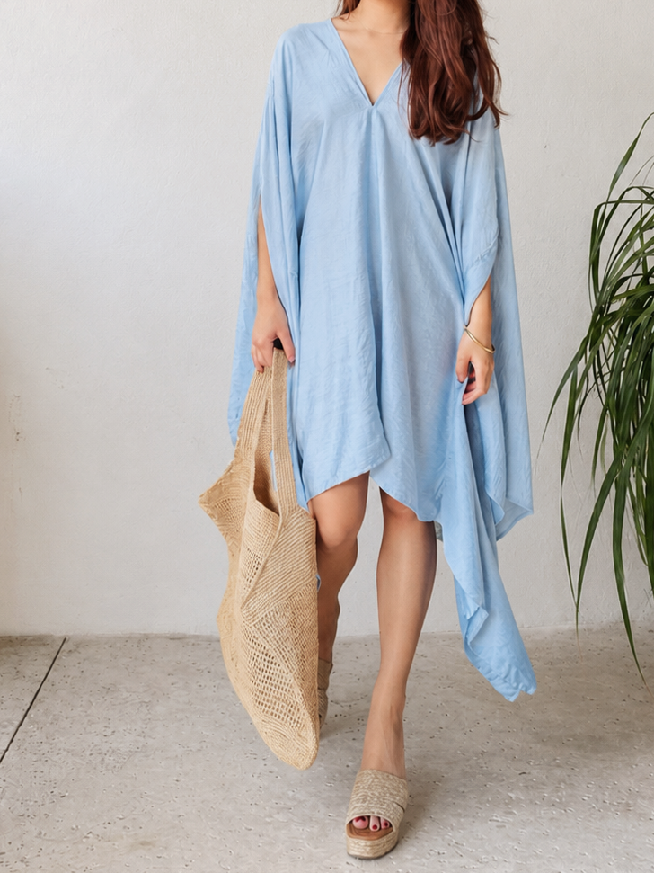 A woman modeling a light blue kaftan dress with a V-neckline and wide draped sleeves, standing against a neutral light gray background. Sky blue Soleil Mini Kaftan Cover Up, lightweight mini kaftan dress and beach cover up styled as a resort top for summer vacation