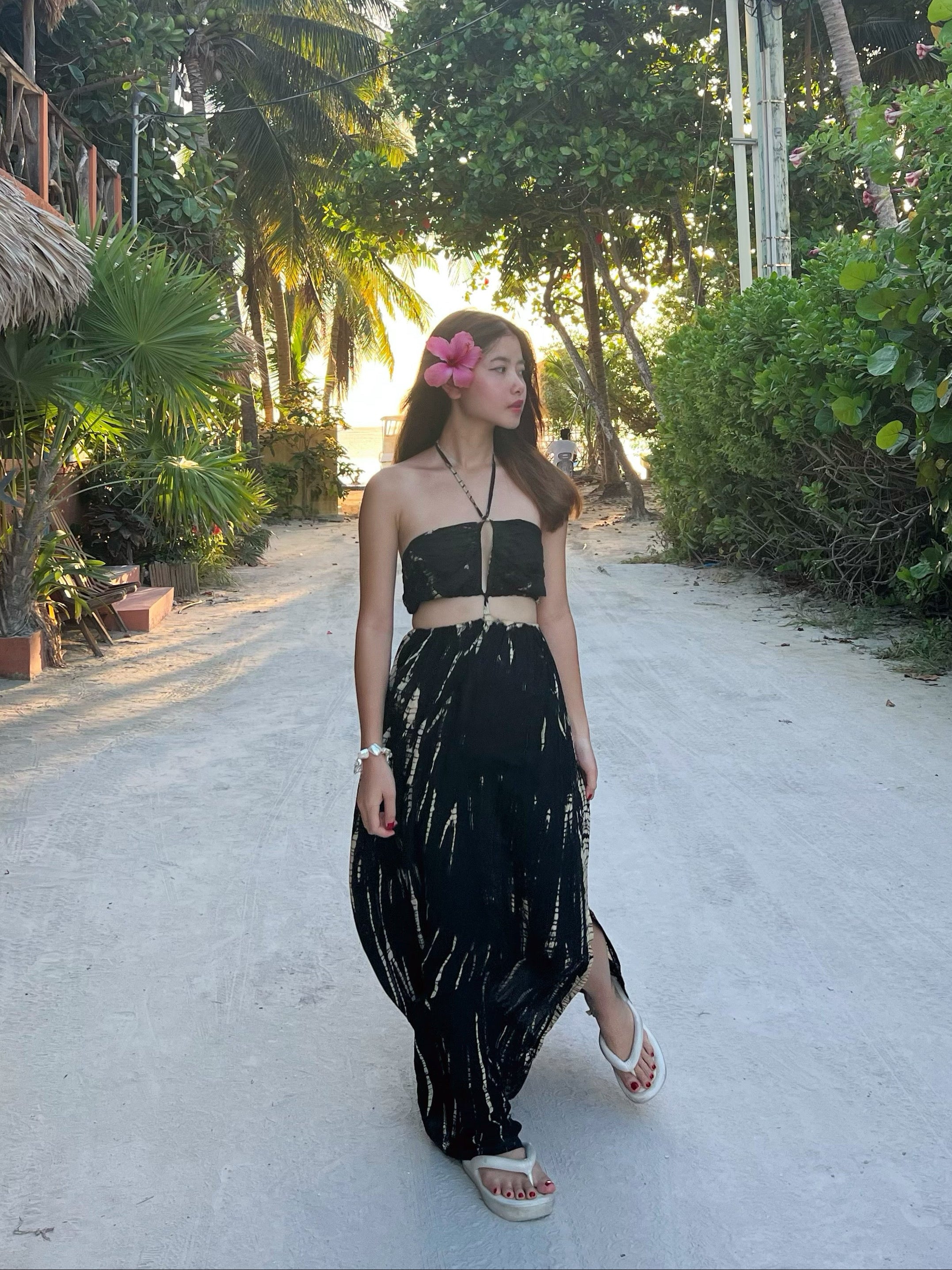 A person wearing a tie-dye cut out maxi dress with a deep v-neckline and spaghetti straps, standing on a pathway with palm trees in the background.