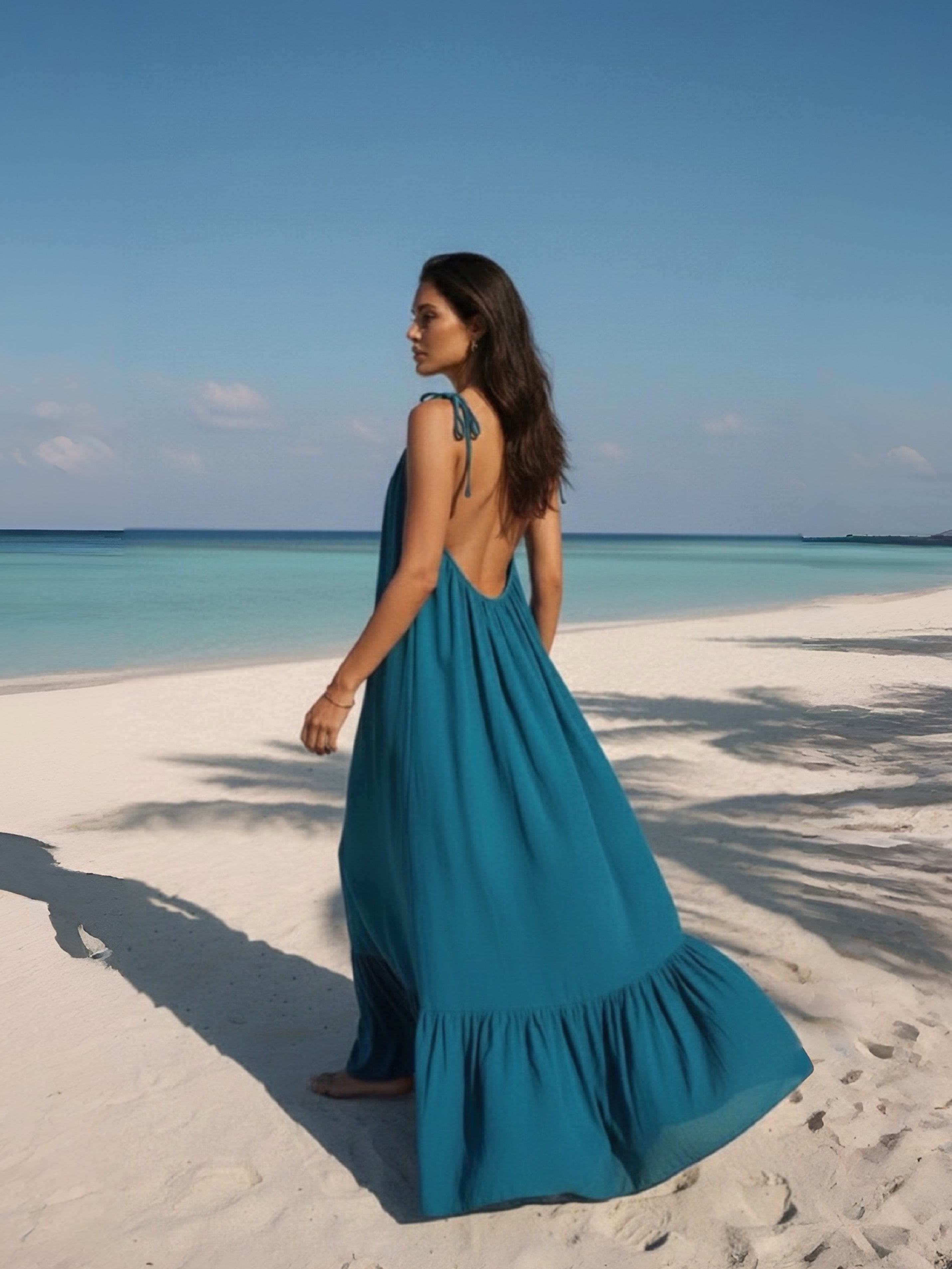 Woman in an open back maxi dress in teal  standing on a sandy beach with clear blue water and sky.