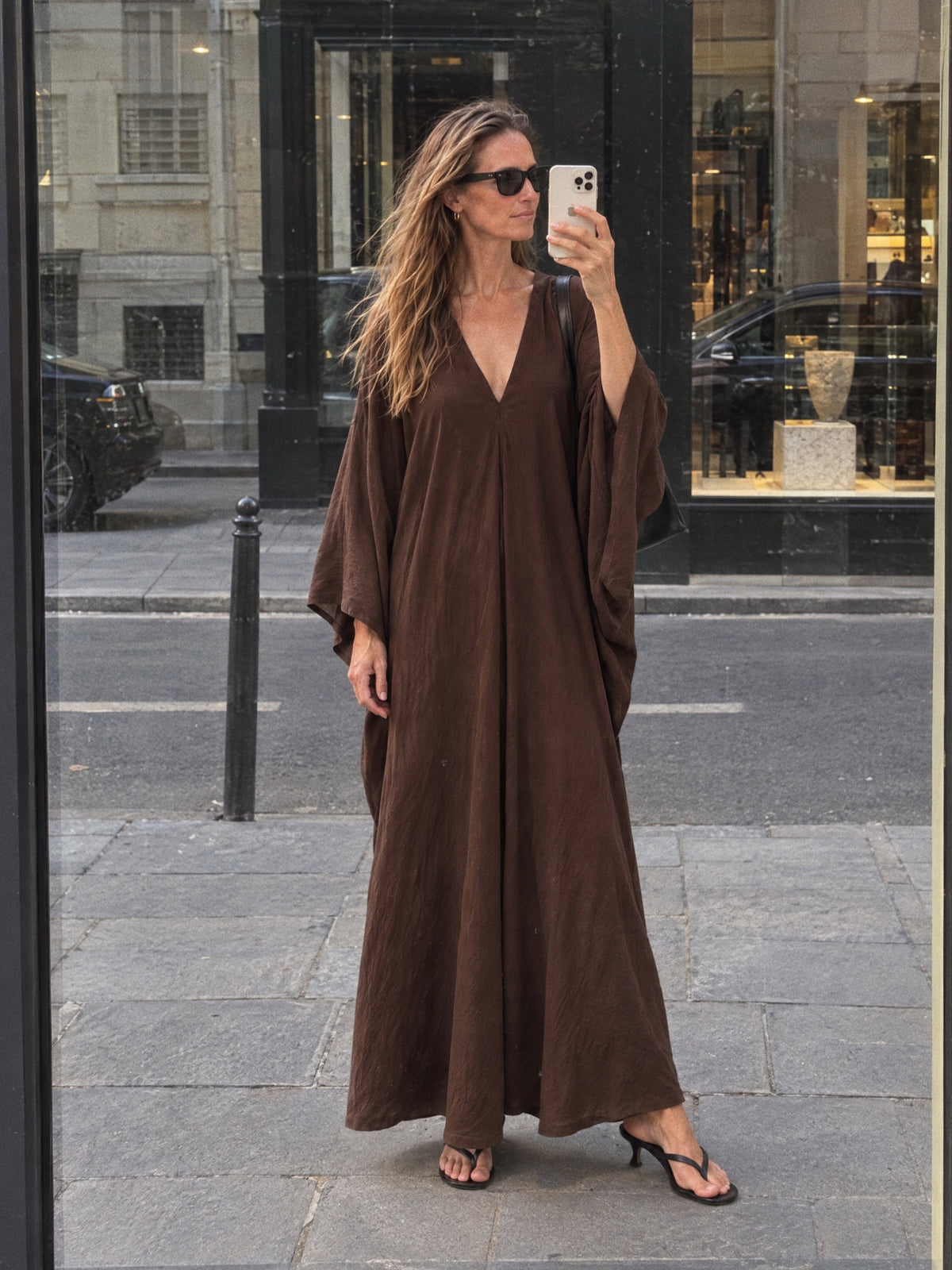 A woman takes a mirror selfie wearing a long, loose-fitting brown maxi kaftan dress with wide draped sleeves and a V-neckline, paired with black heeled sandals on a city sidewalk.