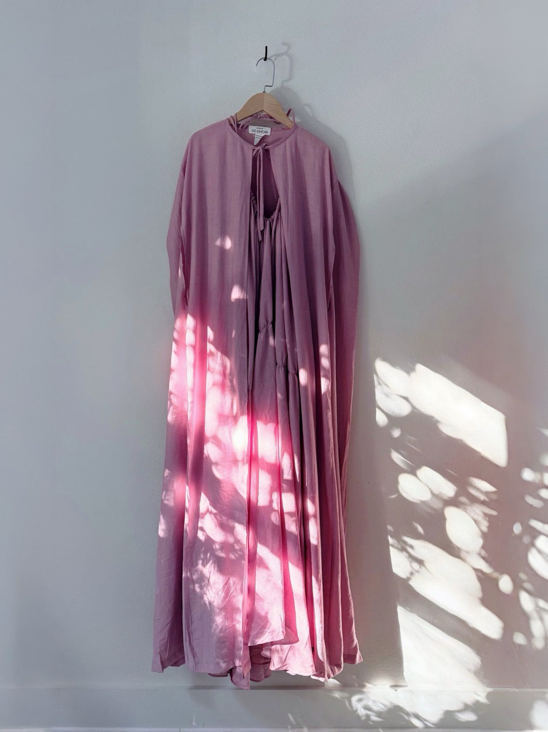 A long-sleeved, high-neck maxi dress in dusty mauve pink hangs on a wooden hanger against a white wall, with dappled sunlight and leaf shadows cast across the garment.