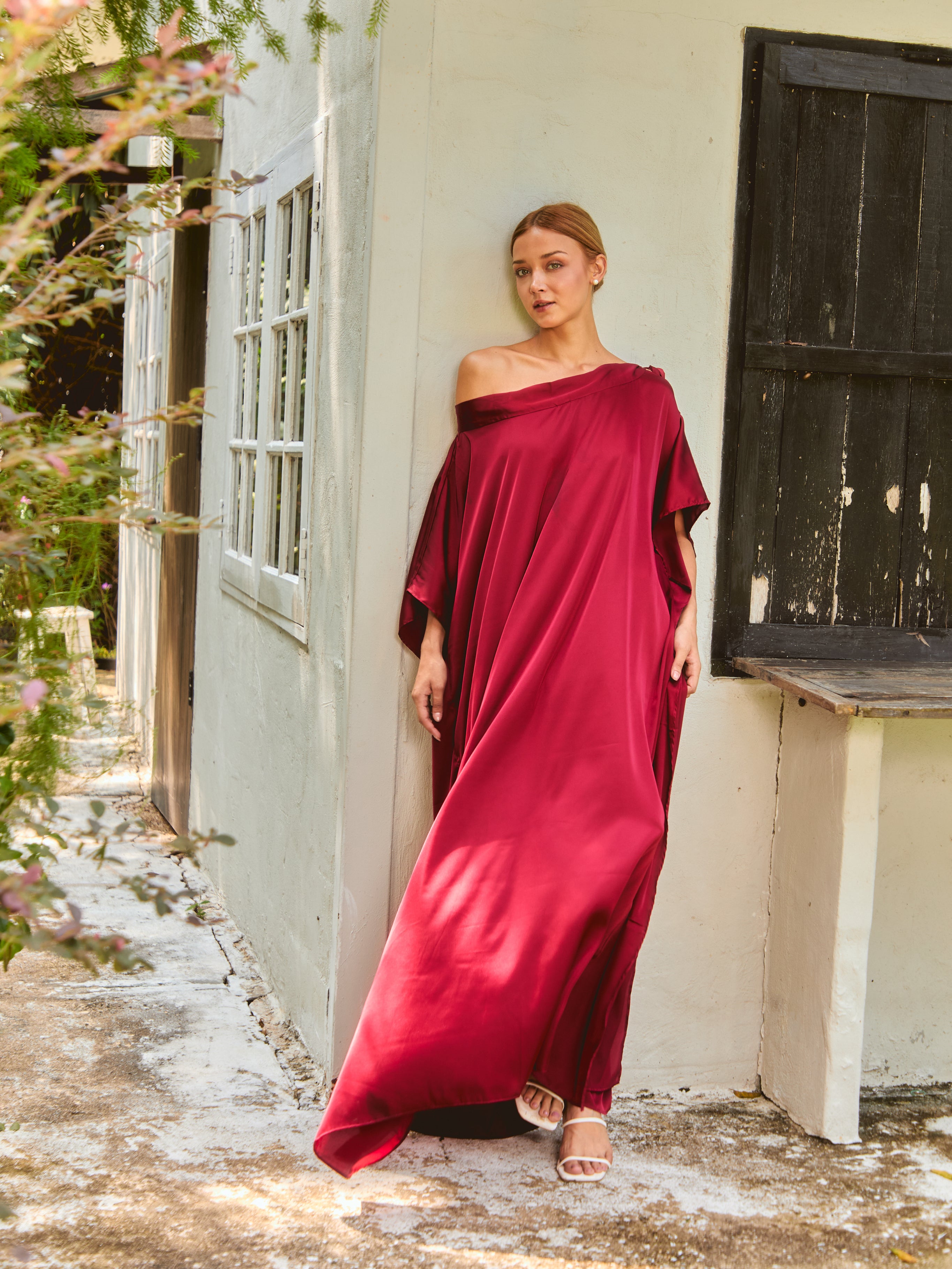 Woman wearing a red off-shoulder  kaftan dress standing in front of a white wall with a wooden door.