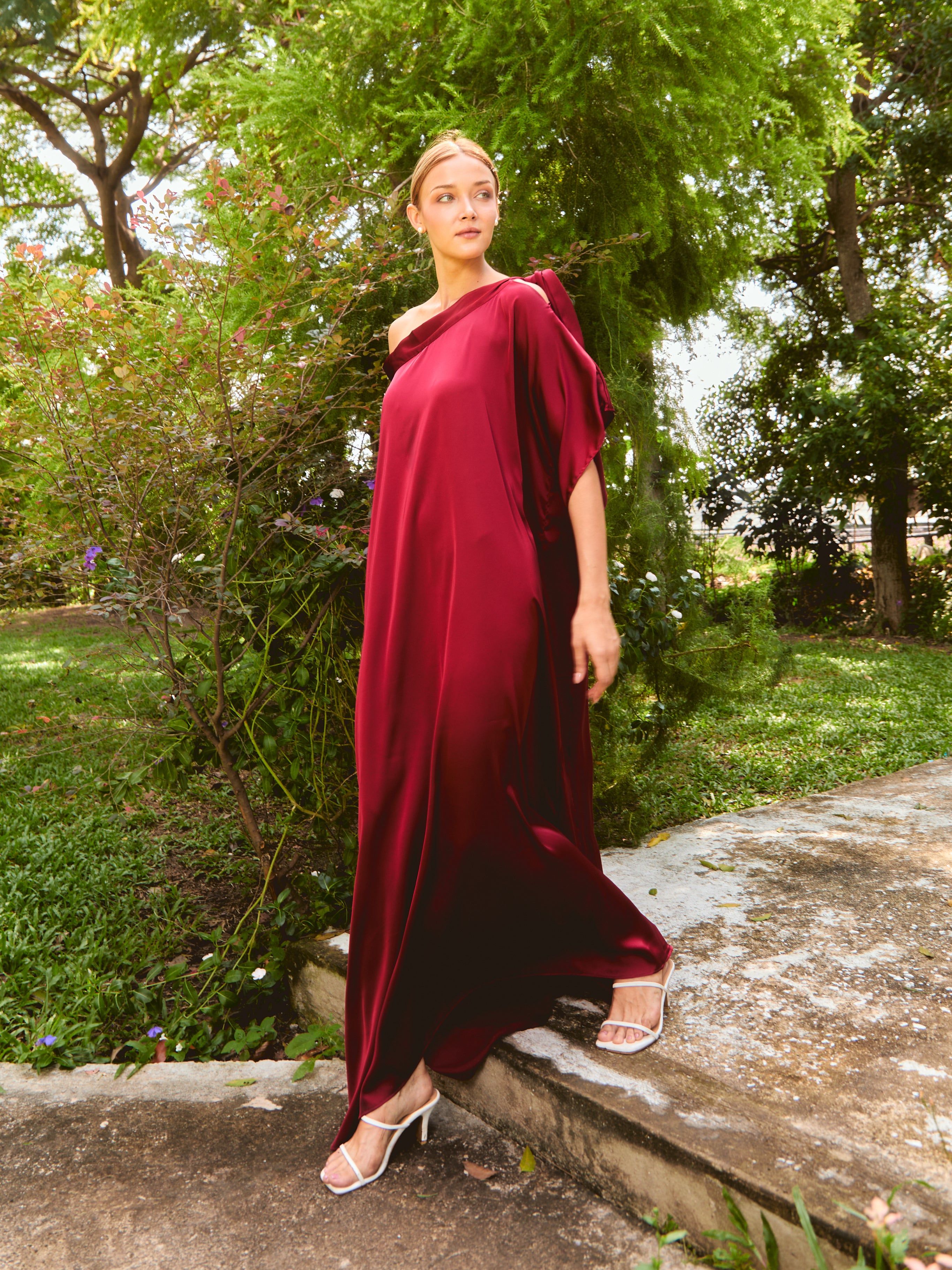 Woman in a burgundy off-shoulder dress standing outdoors near a white wall. The Helena Red Burgundy Kaftan by Coco De Chom features an elegant, free-flowing silhouette with tie details, made from silky eco-friendly satin fabric. Perfect for resort wear, beach vacations, and effortless boho luxury.