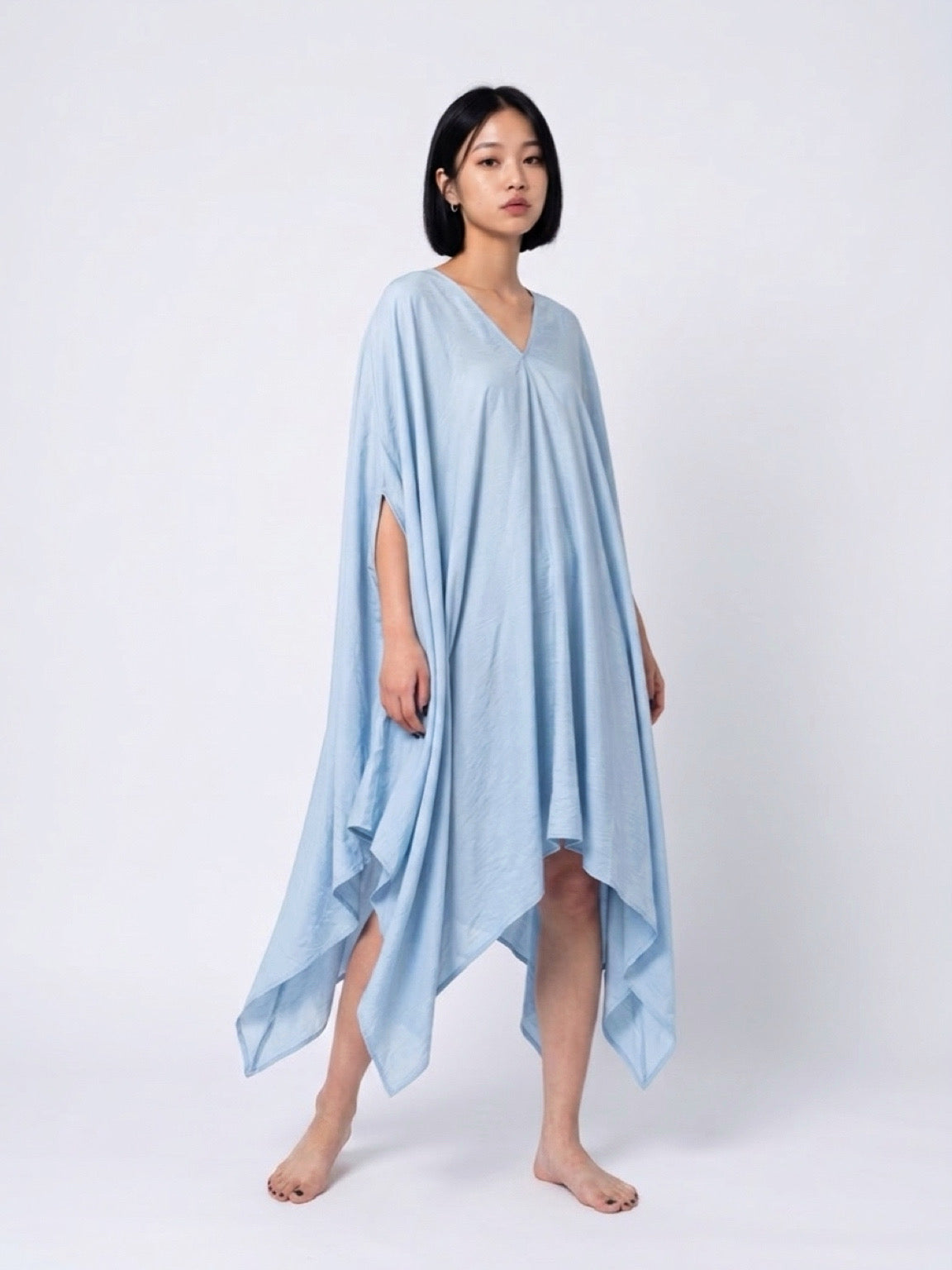 A woman modeling a light blue kaftan dress with a V-neckline and wide draped sleeves, standing against a neutral light gray background. Sky blue Soleil Mini Kaftan Cover Up, lightweight mini kaftan dress and beach cover up styled as a resort top for summer vacation
