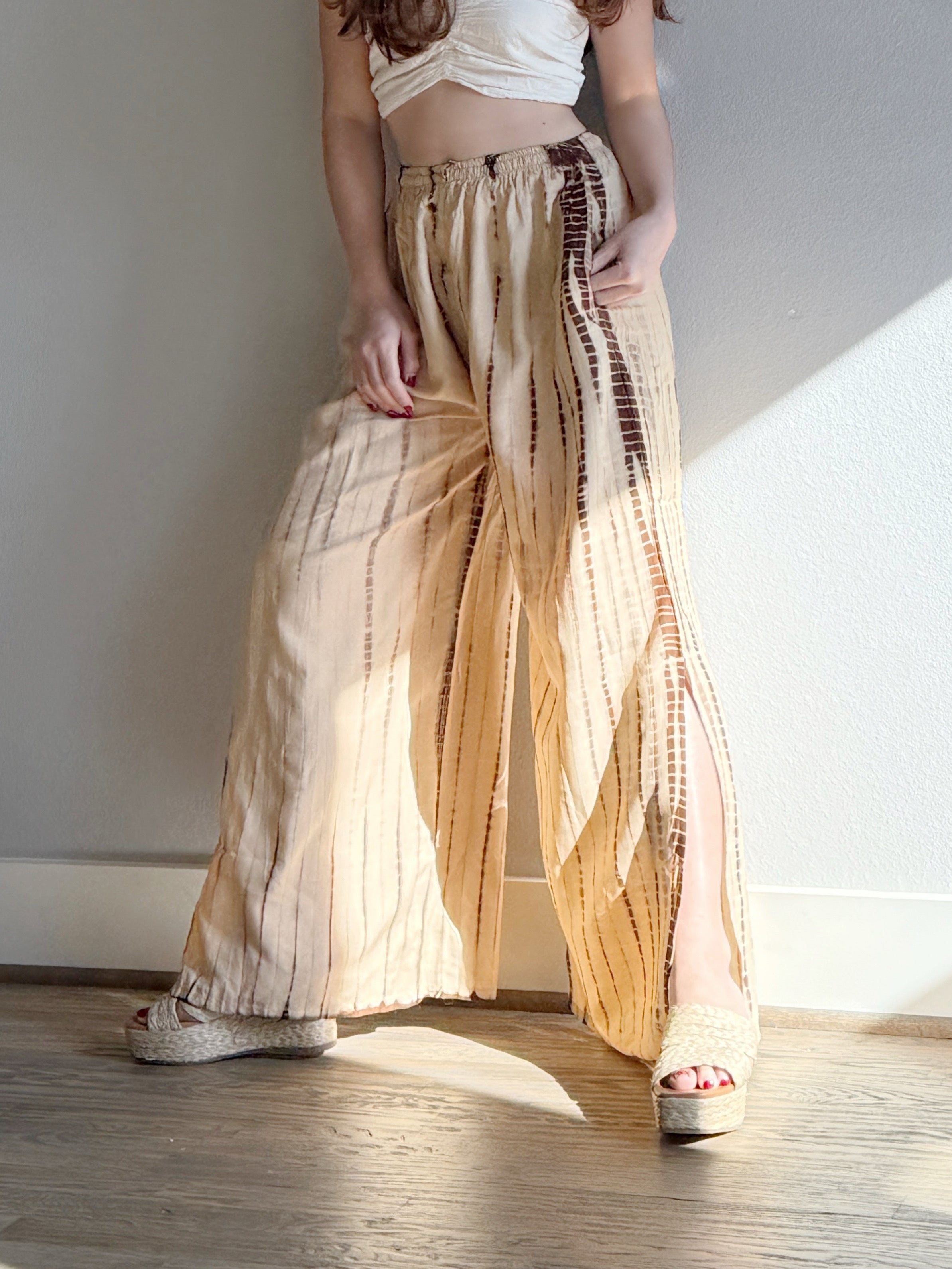 Woman wearing a white top and beige tie dye wide-leg pants, giving a look boho vibe perfect outfit for beach vacation, standing in a room with sunlight streaming in.