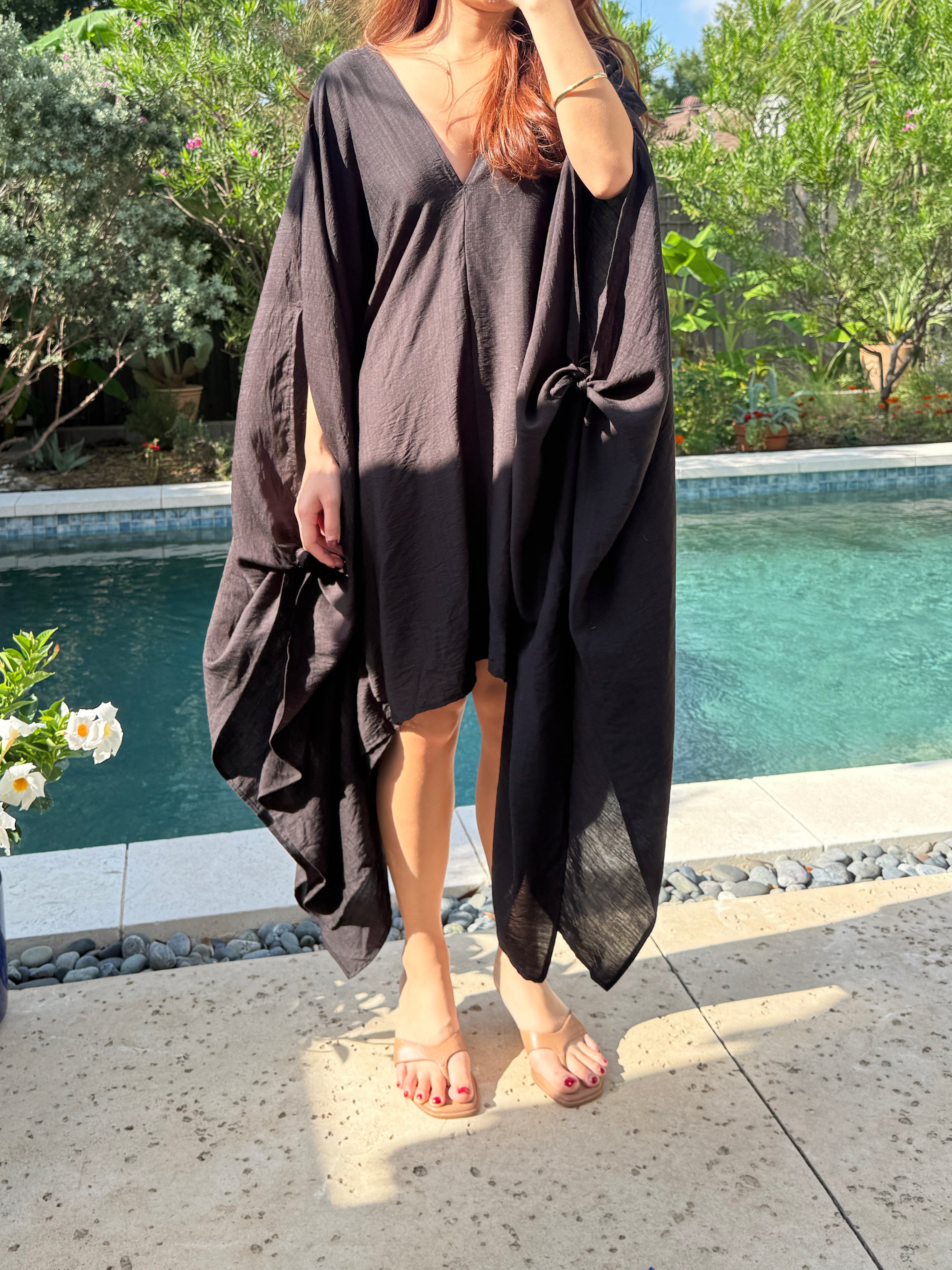 A woman wearing a Black Mini Kaftan Cover Up, oversized beach cover up and petite mini dress with deep V neckline for resort wear and poolside vacation outfits stands by a swimming pool under a white patio umbrella, with green foliage in the background.
