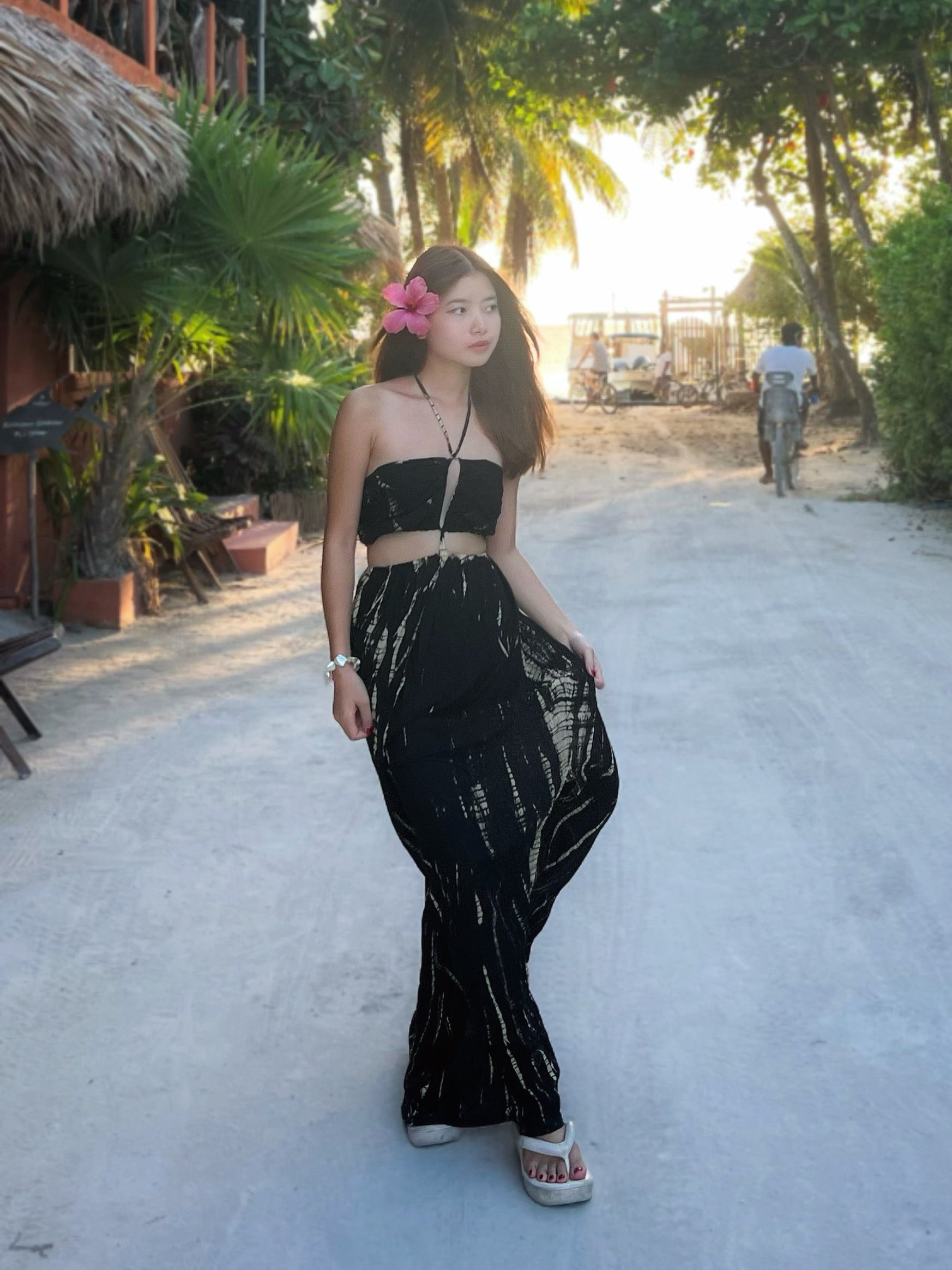 A person wearing a tie-dye cut out maxi dress with a deep v-neckline and spaghetti straps, standing on a pathway with palm trees in the background.