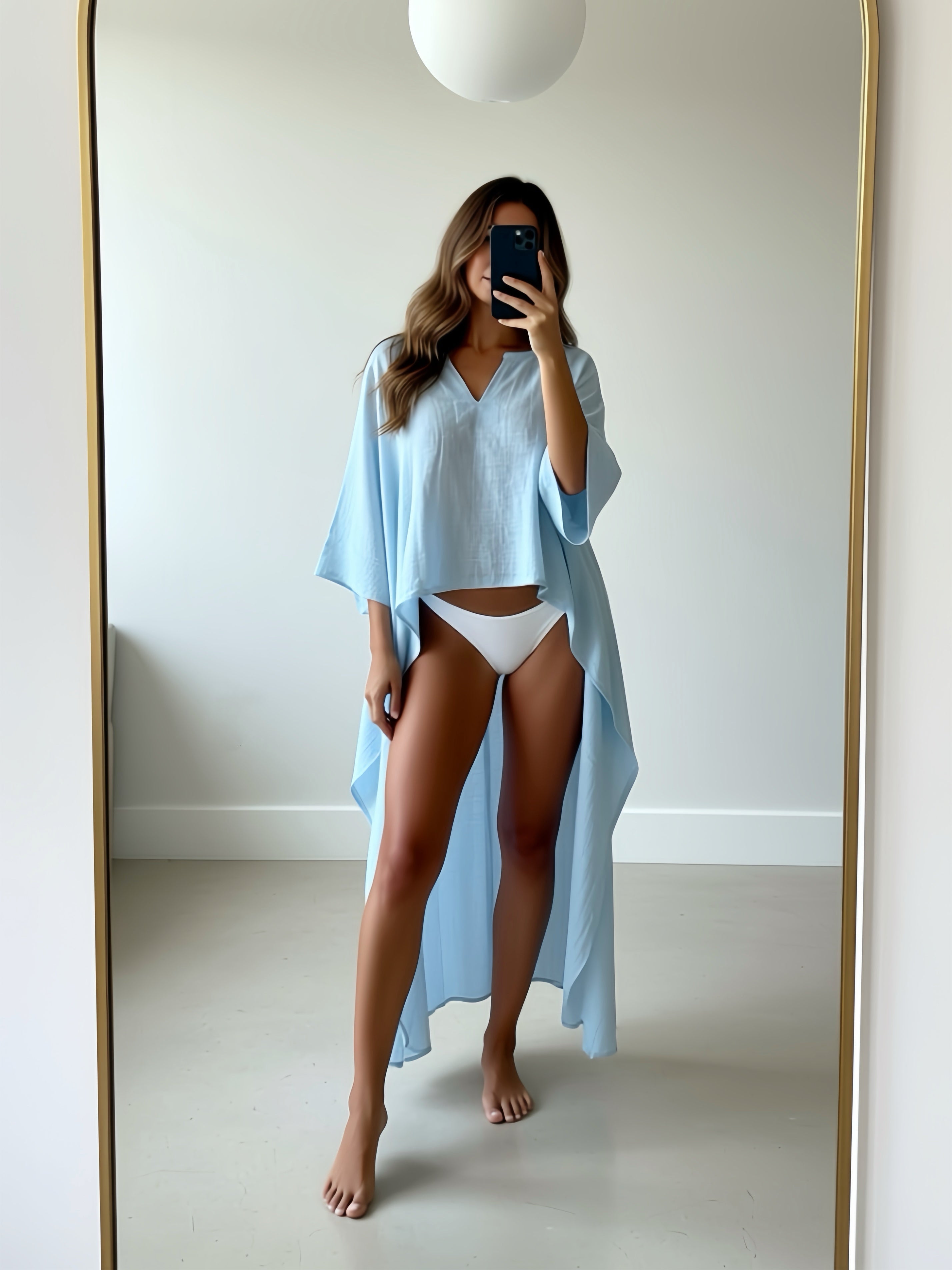 Woman standing on a rocky outcrop by the ocean wearing cotton linen kaftan top light blue worn as beach coverup relaxed fit COCO DE CHOM