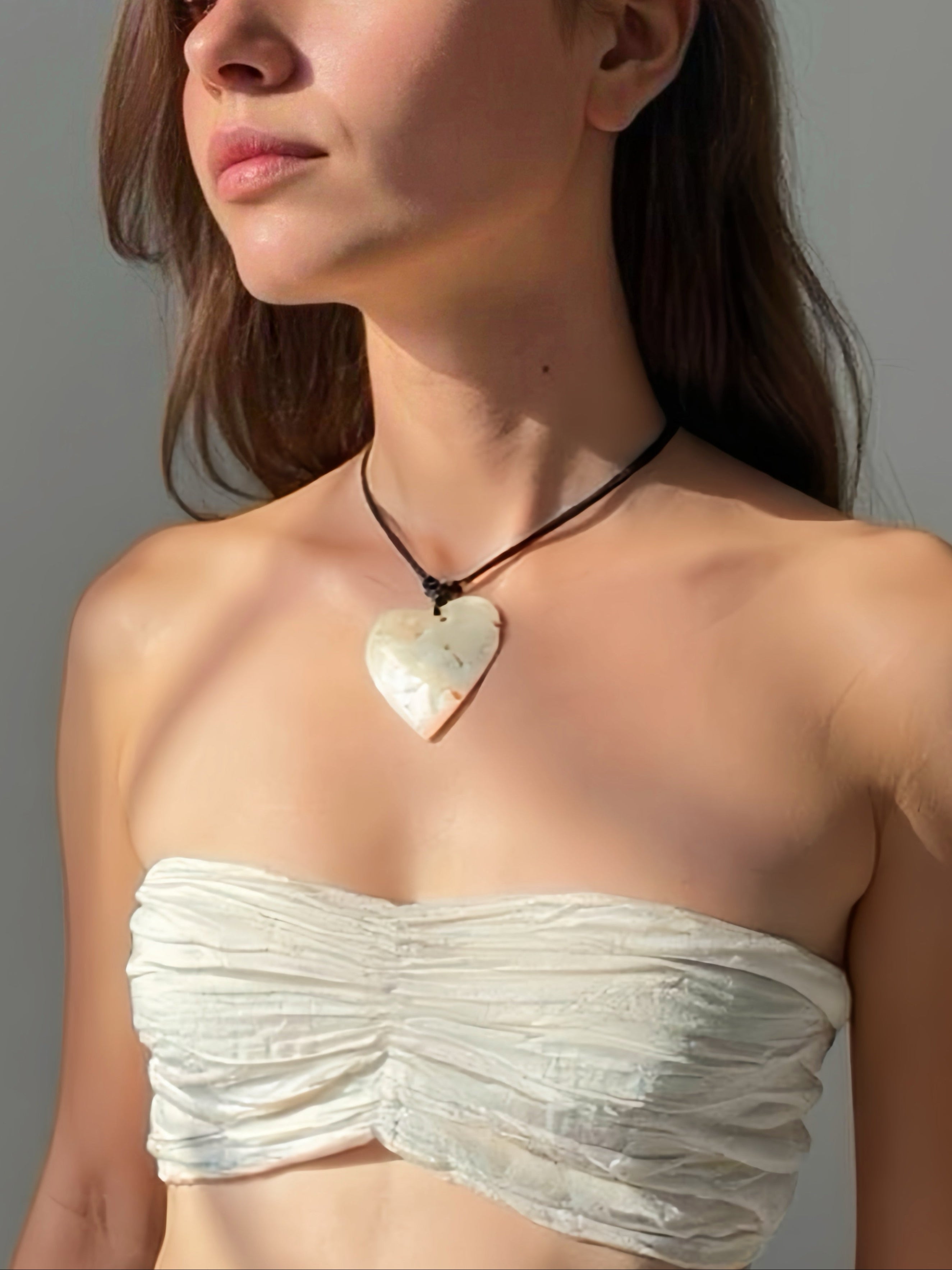 Close-up of a handmade large heart-shaped mother of pearl pendant on an adjustable wax-coated cotton cord, styled as a choker necklace on a woman in natural sunlight. Boho beach jewelry, ethically made 