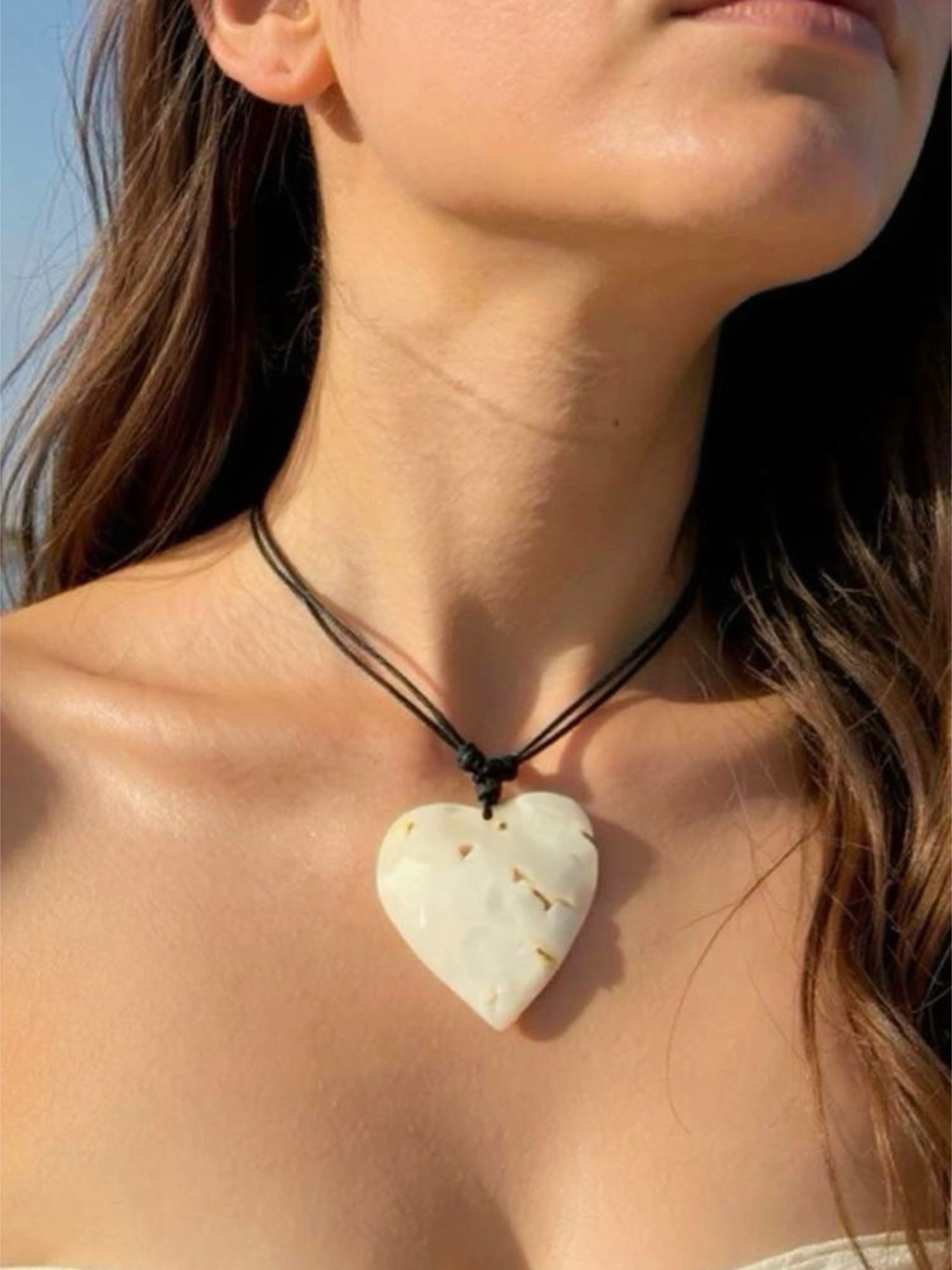 Close-up of a handmade large heart-shaped mother of pearl pendant on an adjustable wax-coated cotton cord, styled as a choker necklace on a woman in natural sunlight. Boho beach jewelry, ethically made 