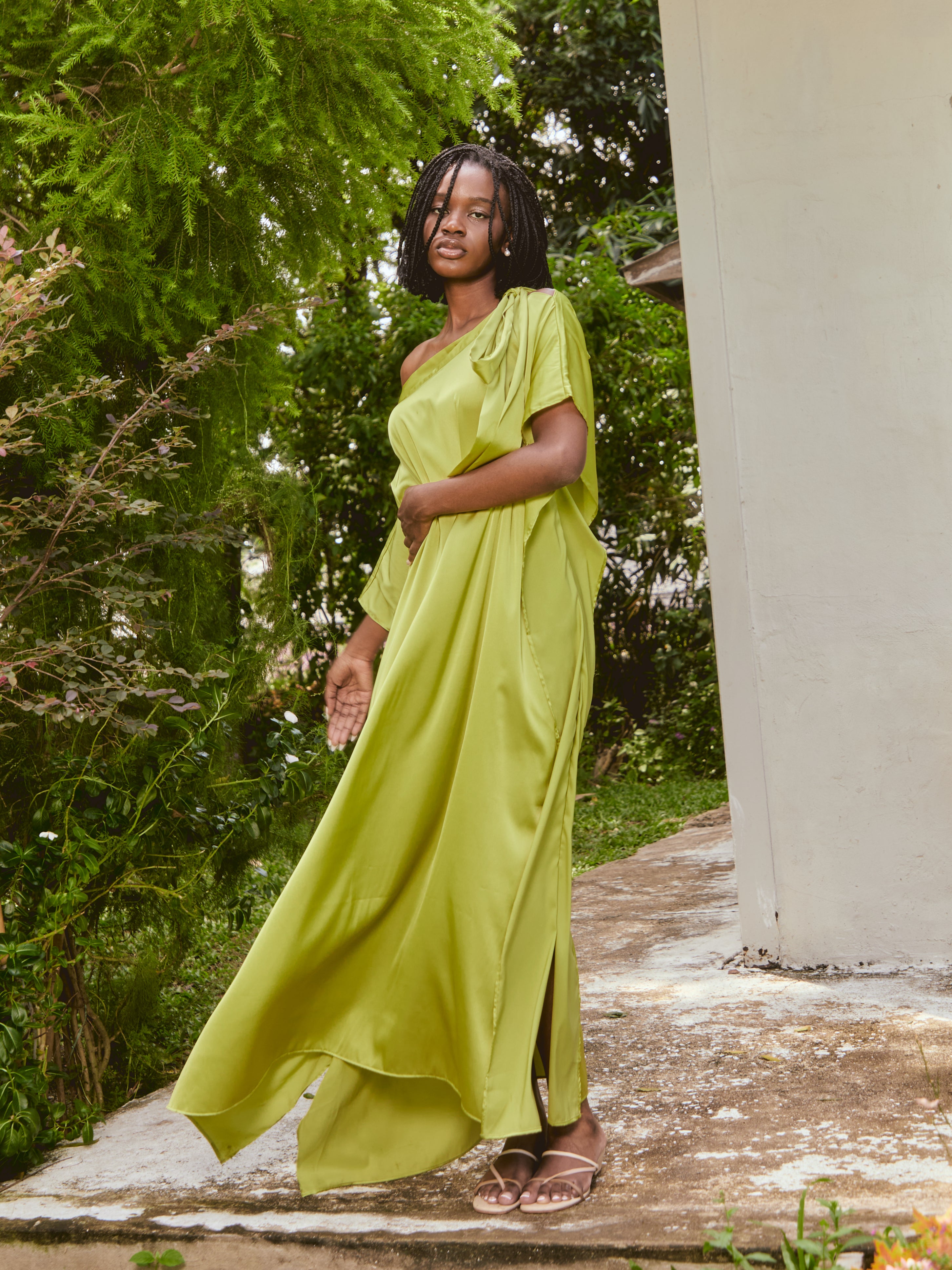 Woman in a lemon green kaftan dress standing on grass with a house and tree in the background. The Helena Lemon Green Kaftan by Coco De Chom is an off-shoulder satin resort dress with a free-flowing silhouette, tie details, and silky eco-friendly fabric — perfect for beach vacations, resort wear, and effortless boho luxury.