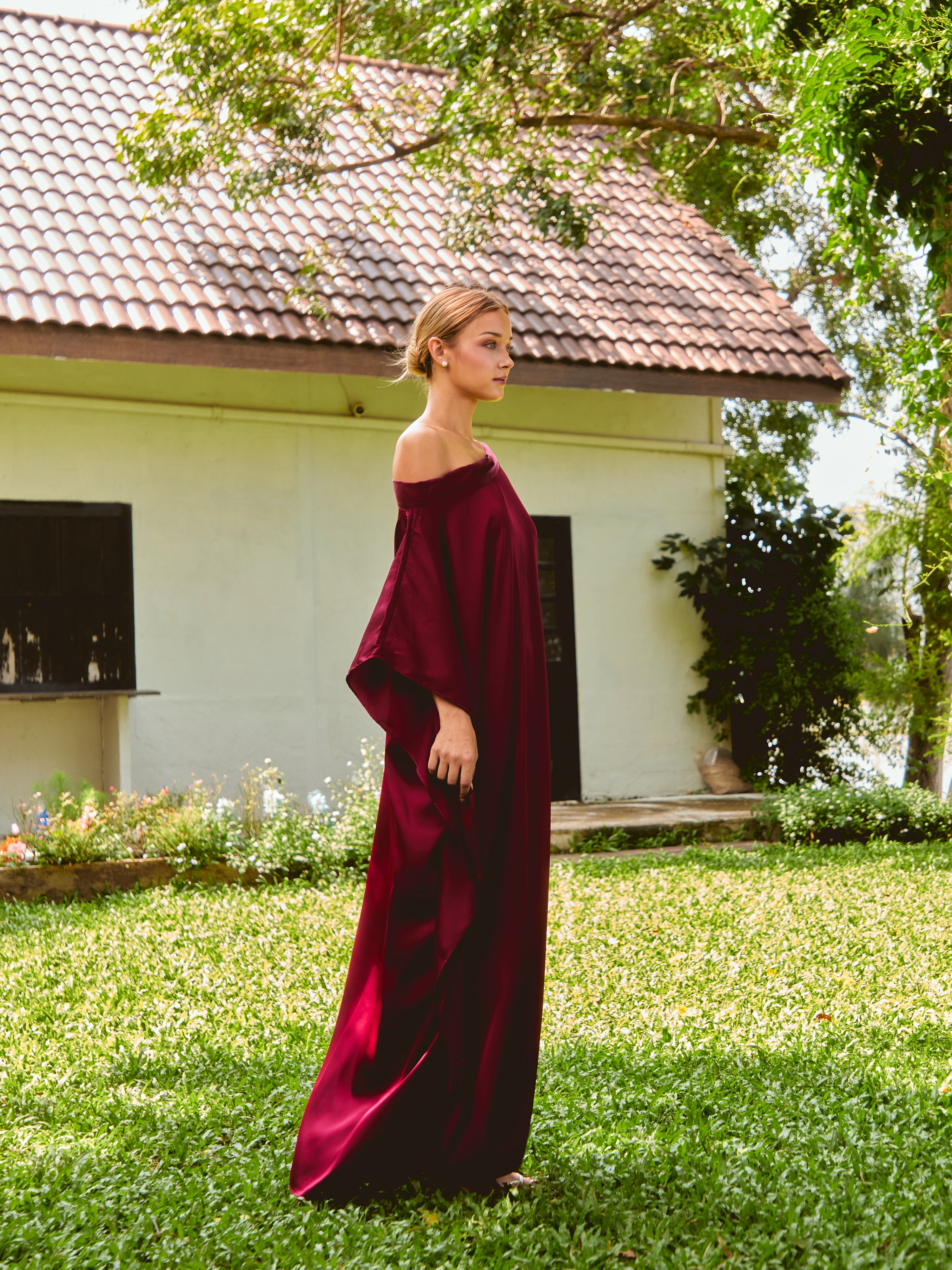 Woman wearing a burgundy off-shoulder dress standing outdoors. The Helena Red Burgundy Kaftan by Coco De Chom features an elegant, free-flowing silhouette with tie details, made from silky eco-friendly satin fabric. Perfect for resort wear, beach vacations, and effortless boho luxury.