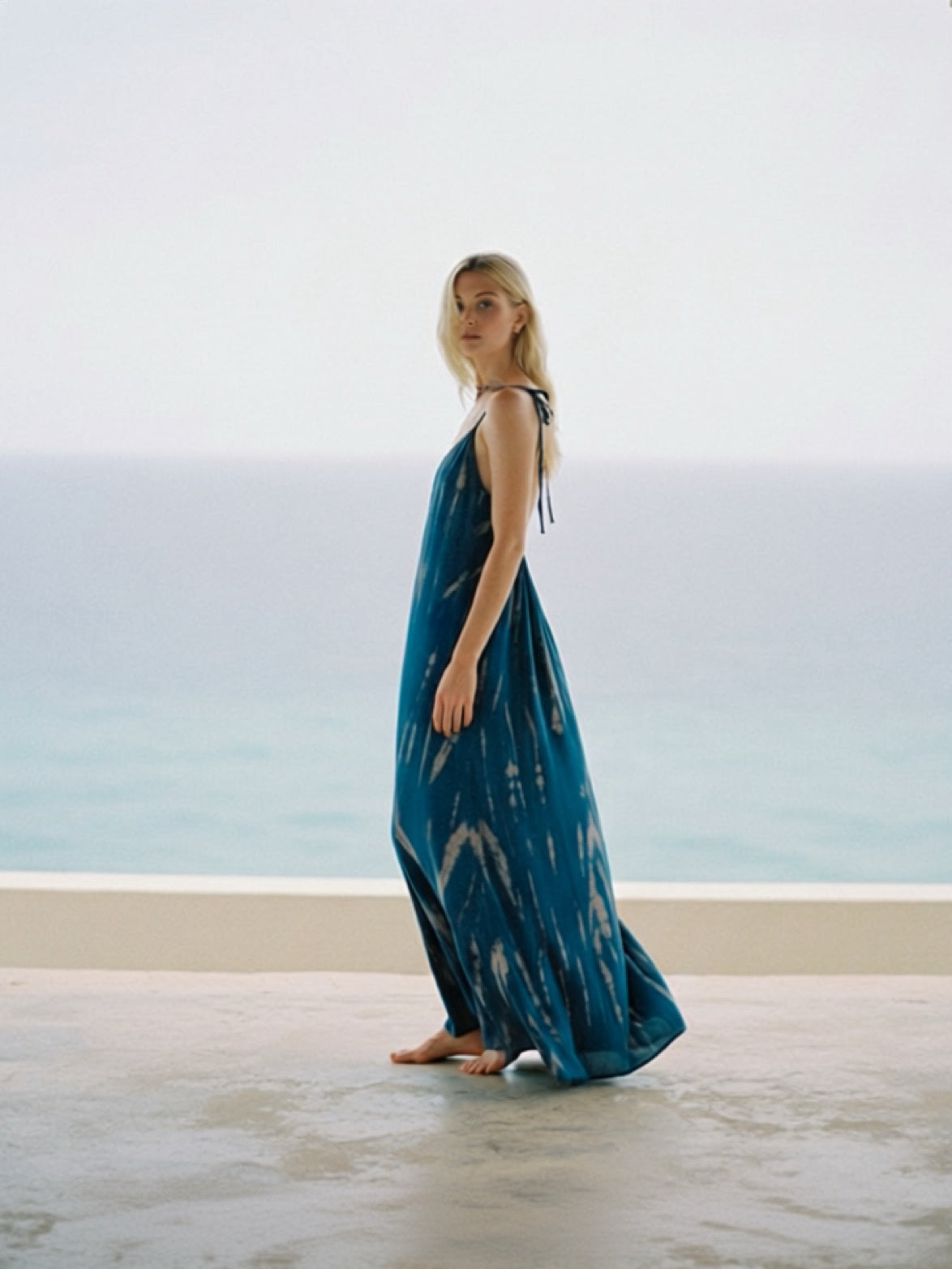 Woman in a teal open back maxi dress standing on a beach with ocean and  resortwear in the background