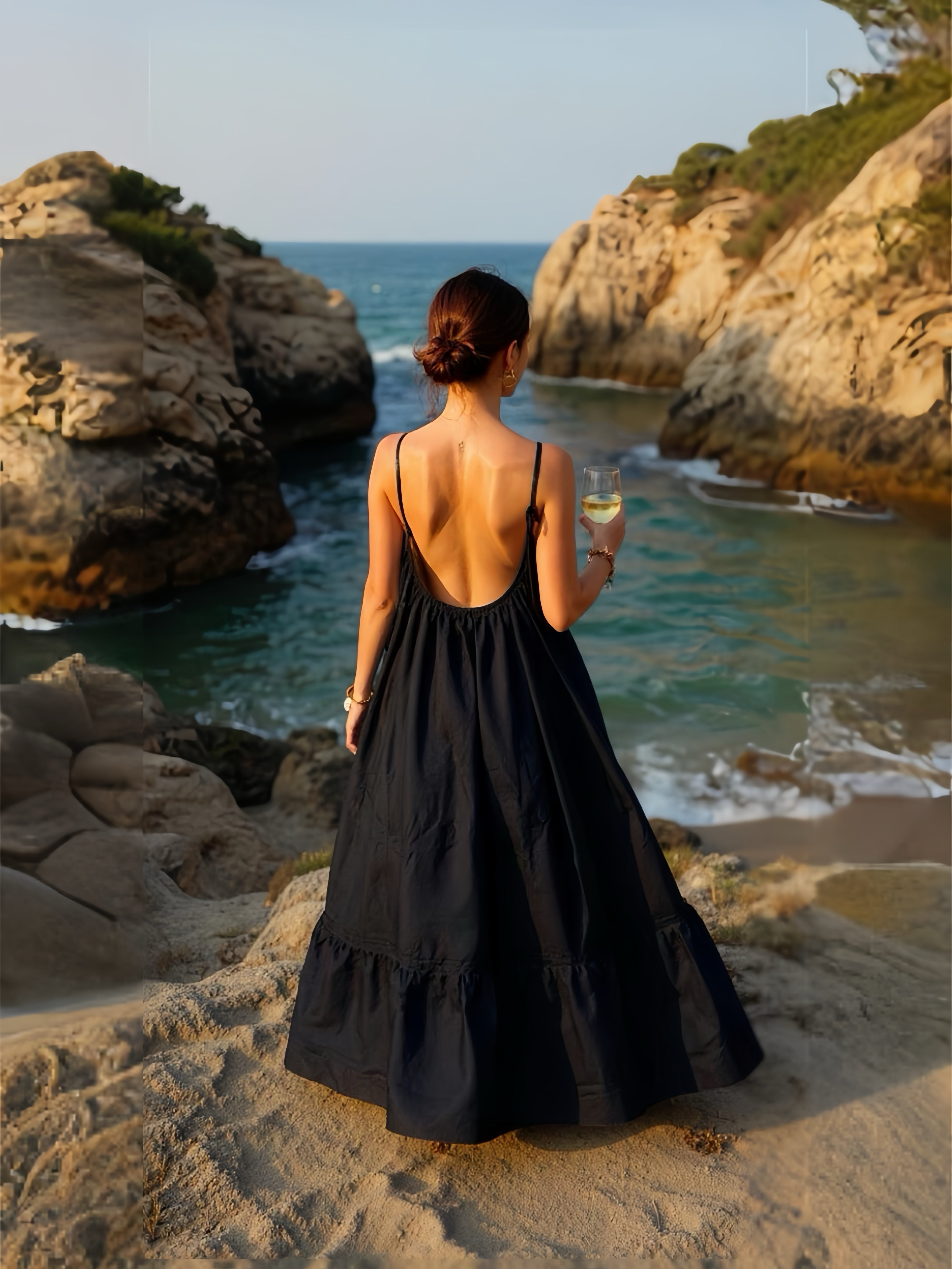 Woman in a hand-dyed black backless maxi dress standing by the sea, holding a glass of wine, wearing boho resortwear by Coco de Chom