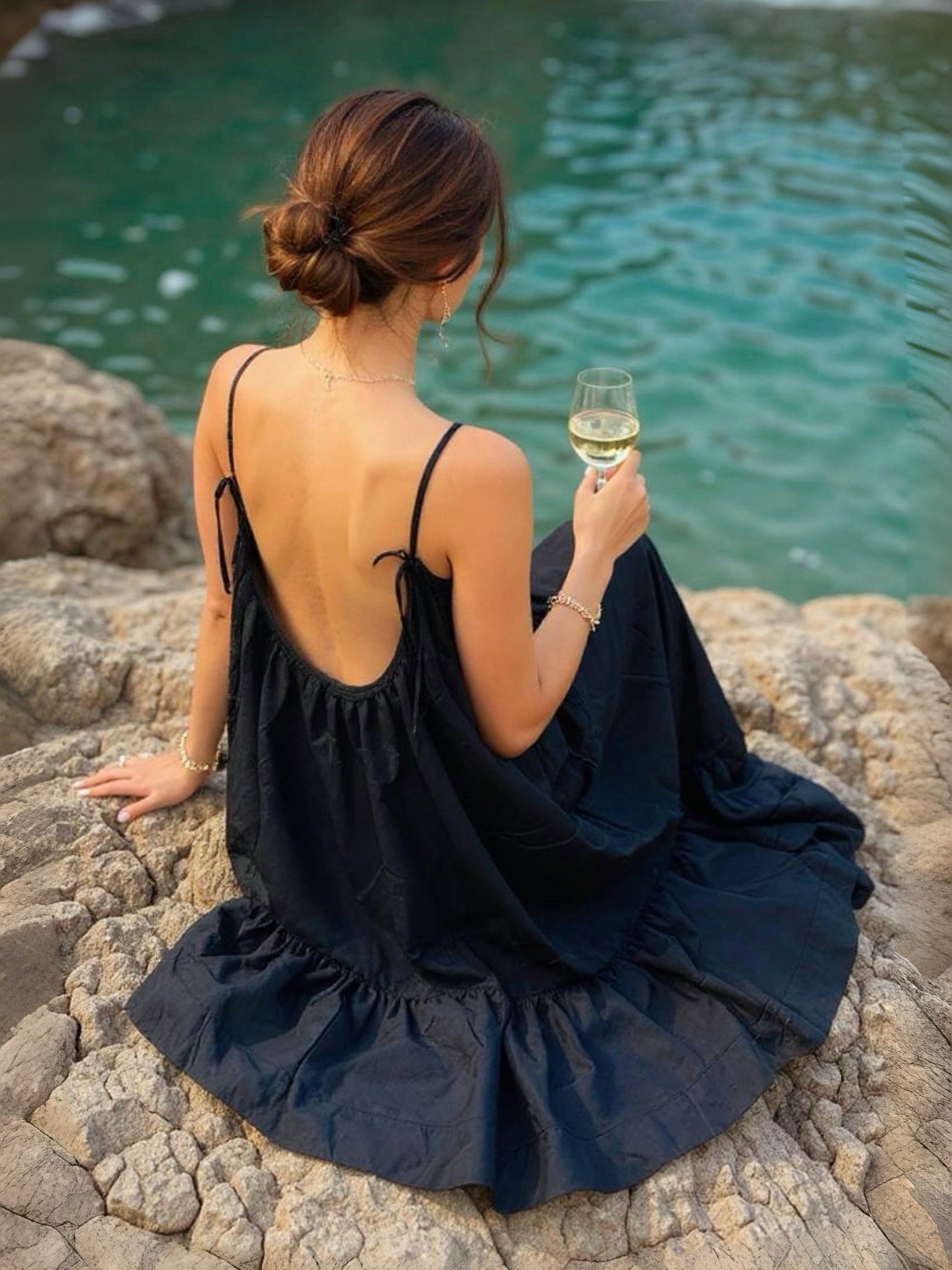 Woman in a hand-dyed black backless maxi dress standing by the sea, holding a glass of wine, wearing boho resortwear by Coco de Chom