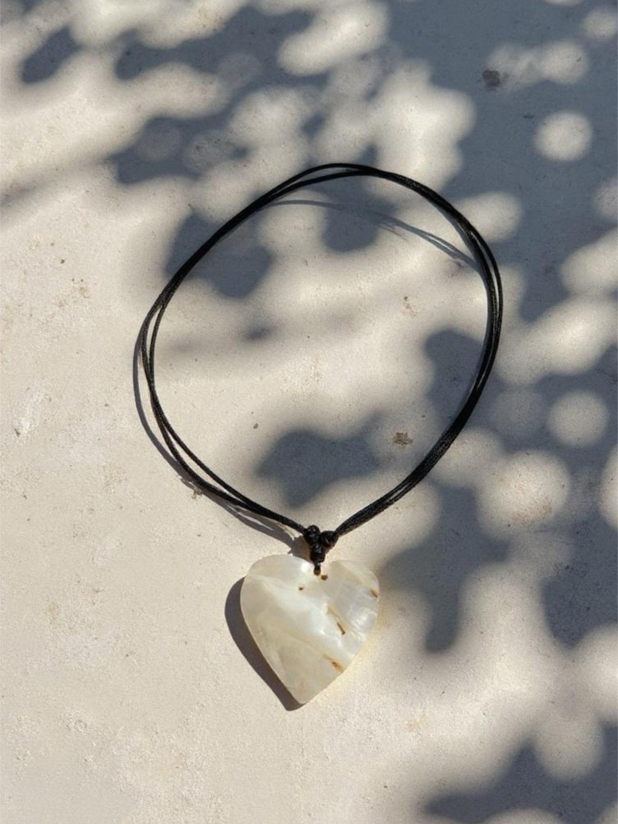 Close-up of a handmade large heart-shaped mother of pearl pendant on an adjustable wax-coated cotton cord, styled as a choker necklace on a woman in natural sunlight. Boho beach jewelry, ethically made 