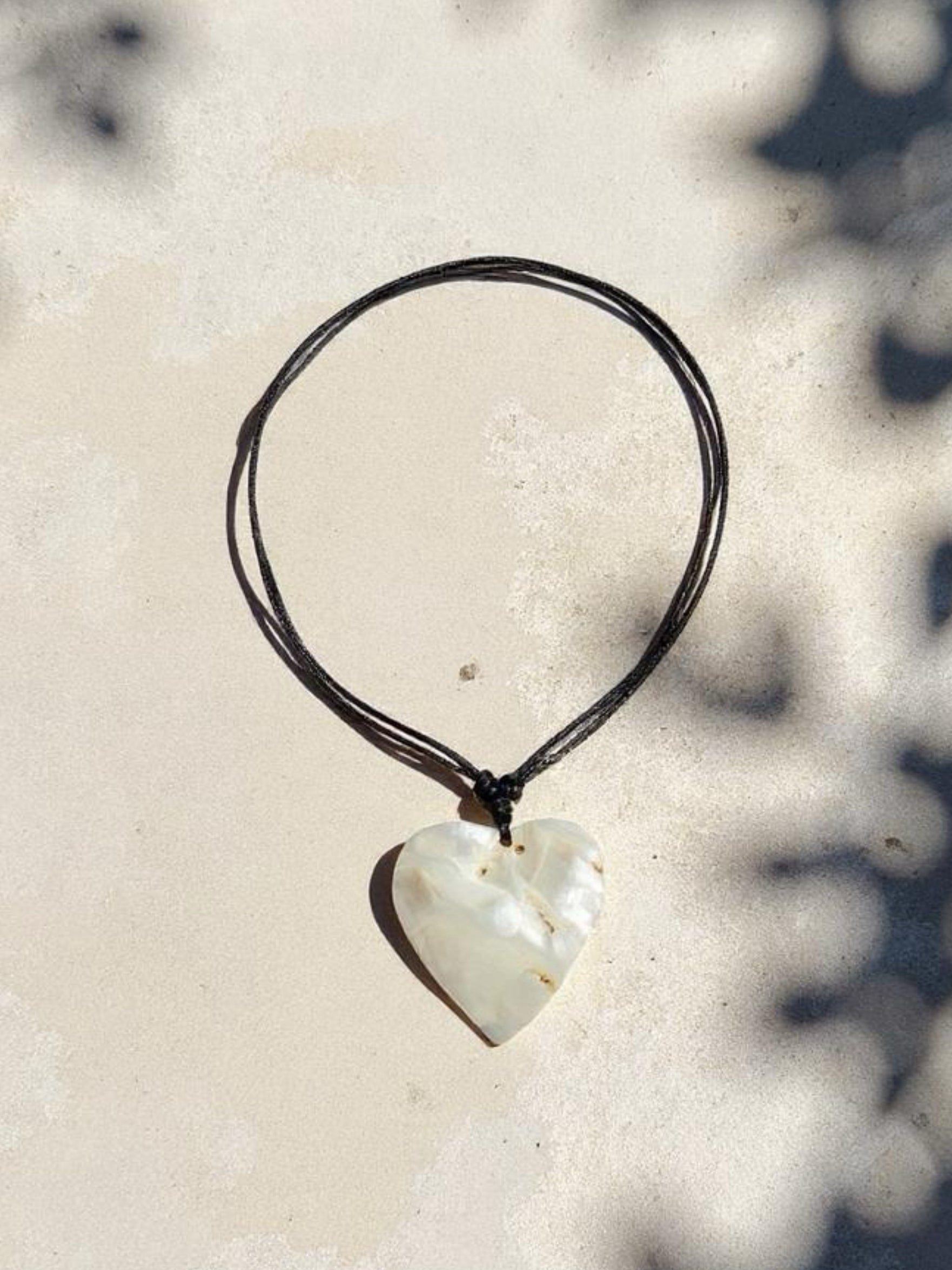Close-up of a handmade large heart-shaped mother of pearl pendant on an adjustable wax-coated cotton cord, styled as a choker necklace on a woman in natural sunlight. Boho beach jewelry, ethically made 