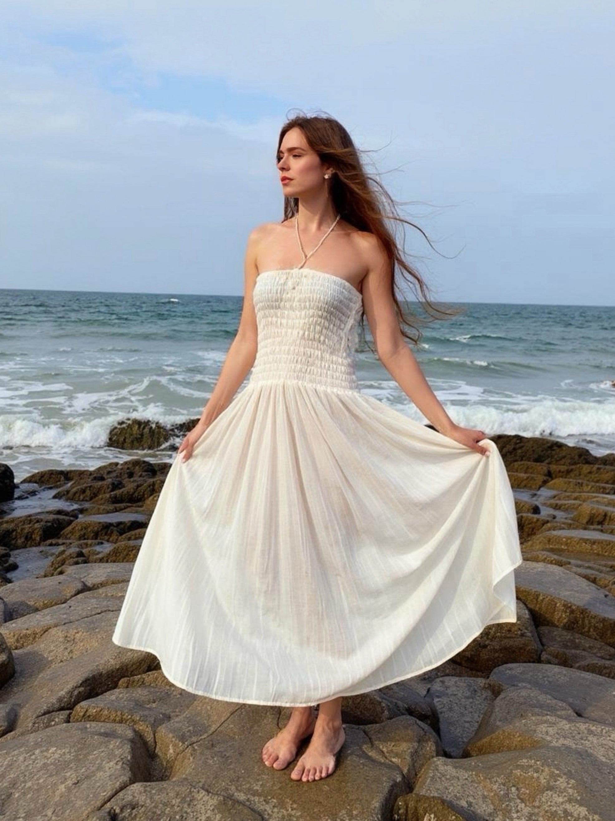 Woman in a white cotton smock dress standing on rocky beach with ocean in the background