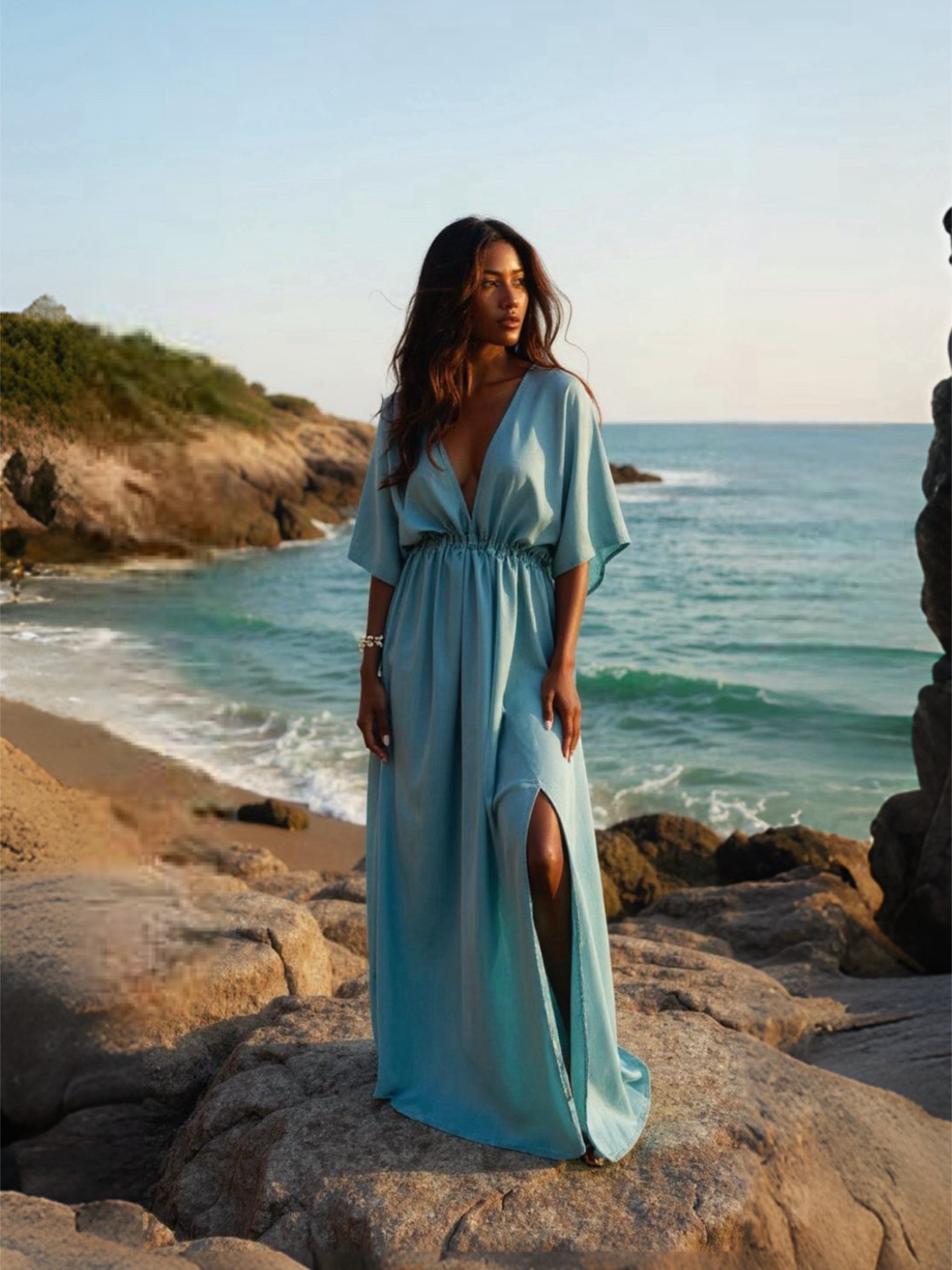 Woman in a light teal kaftan dress standing on a rocky beach with ocean and sky in the background
