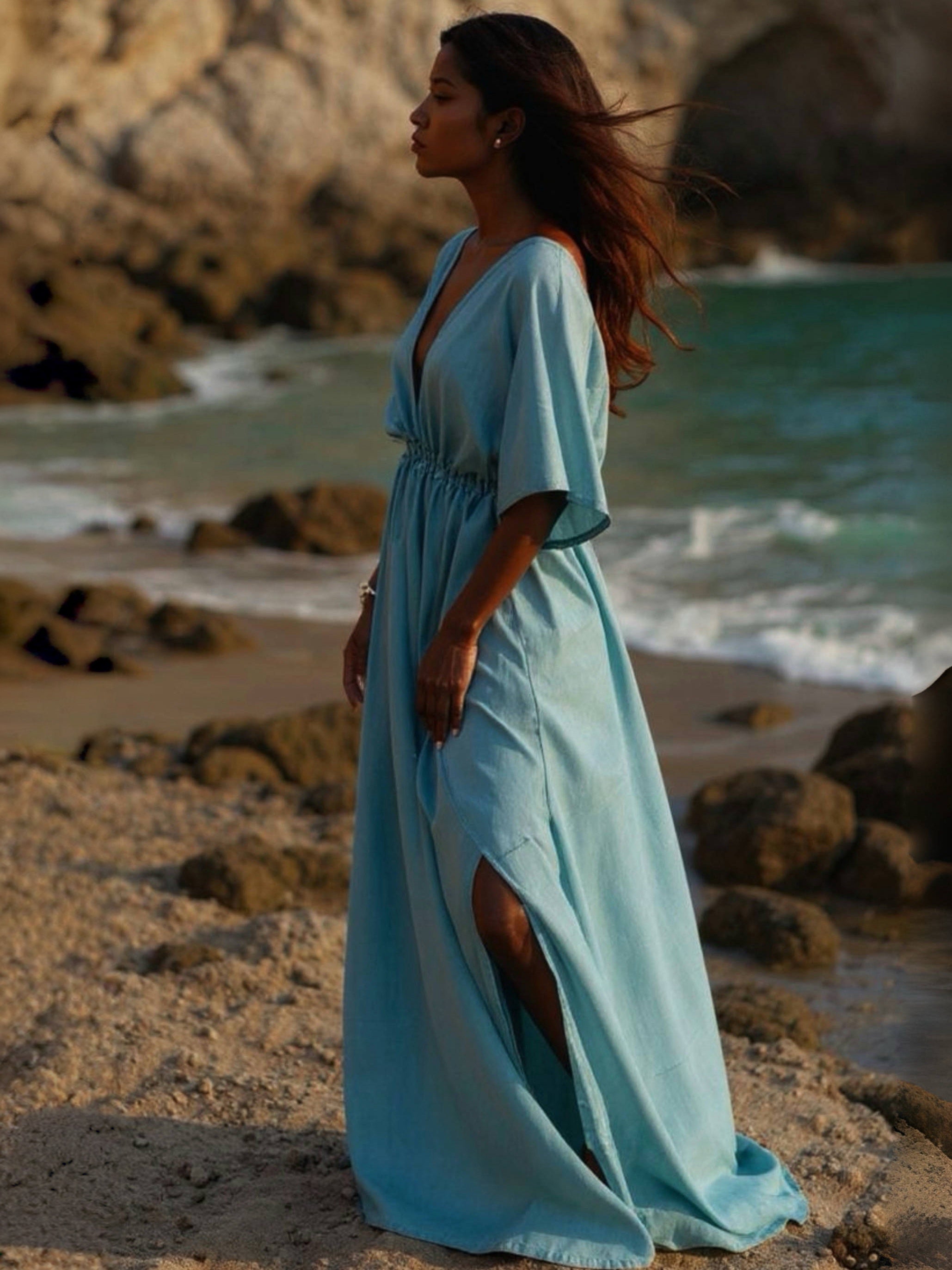 Woman in a light blue goddess maxi dress standing on a beach with waves and rocks in the background