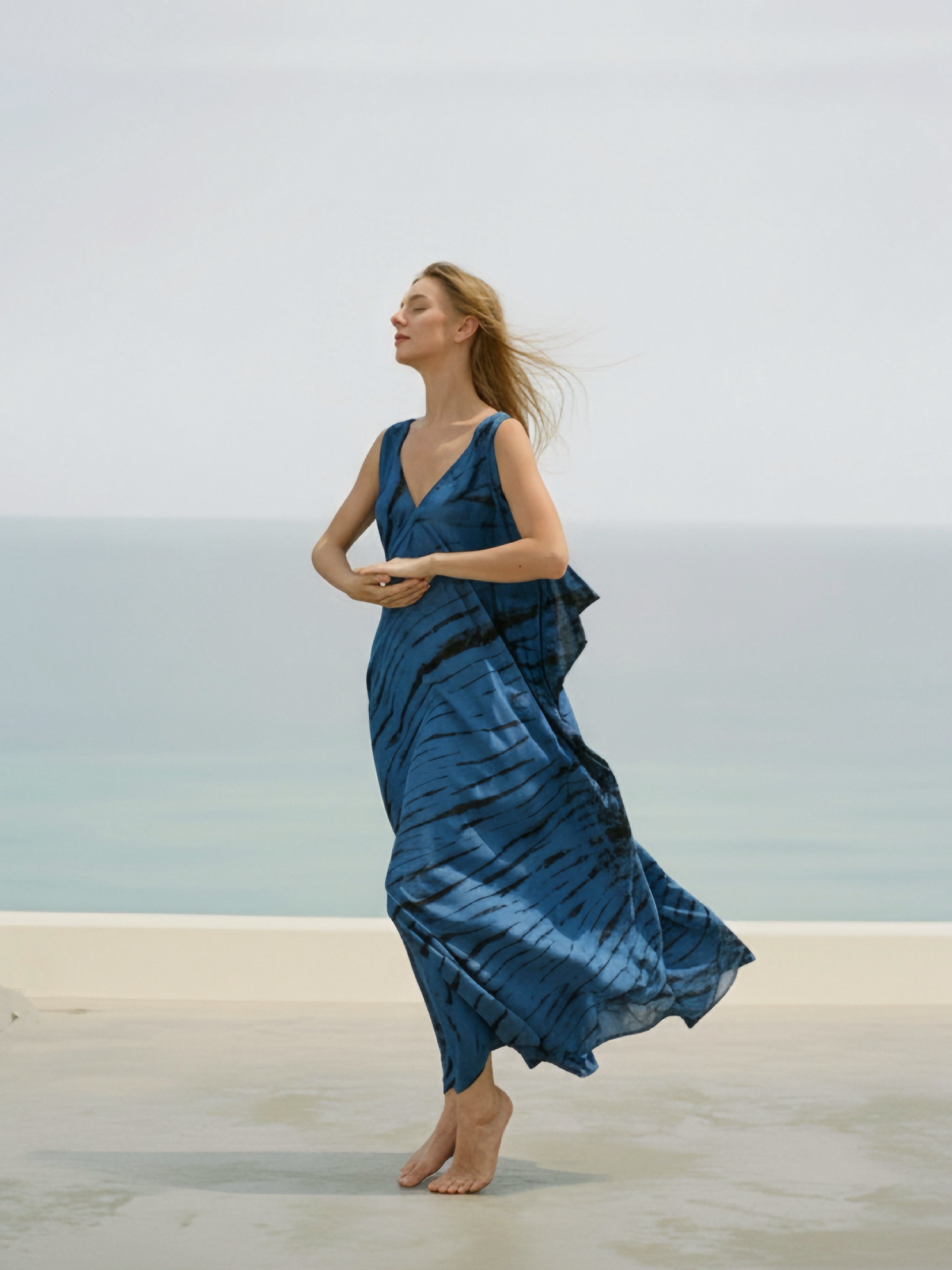 Woman wearing a tie-dye V-neck kaftan dress, a blue boho oversized caftan, resort dress - bohemian long kaftan for beach resortwear from Coco De Chom, standing on a beach with the ocean in the background.