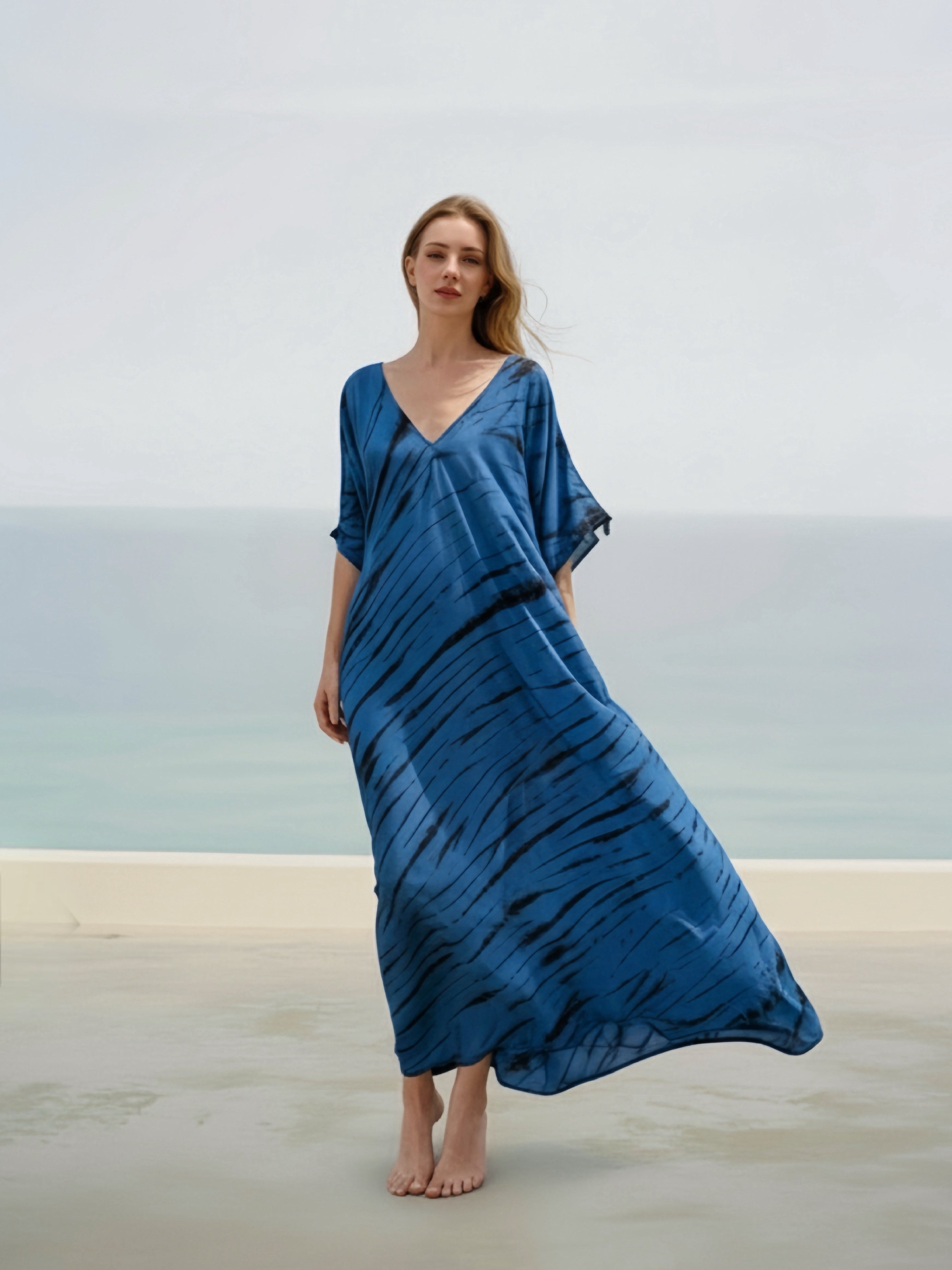 Woman wearing a tie-dye V-neck kaftan dress, a blue boho oversized caftan, resort dress - bohemian long kaftan for beach resortwear from Coco De Chom, standing on a beach with the ocean in the background.