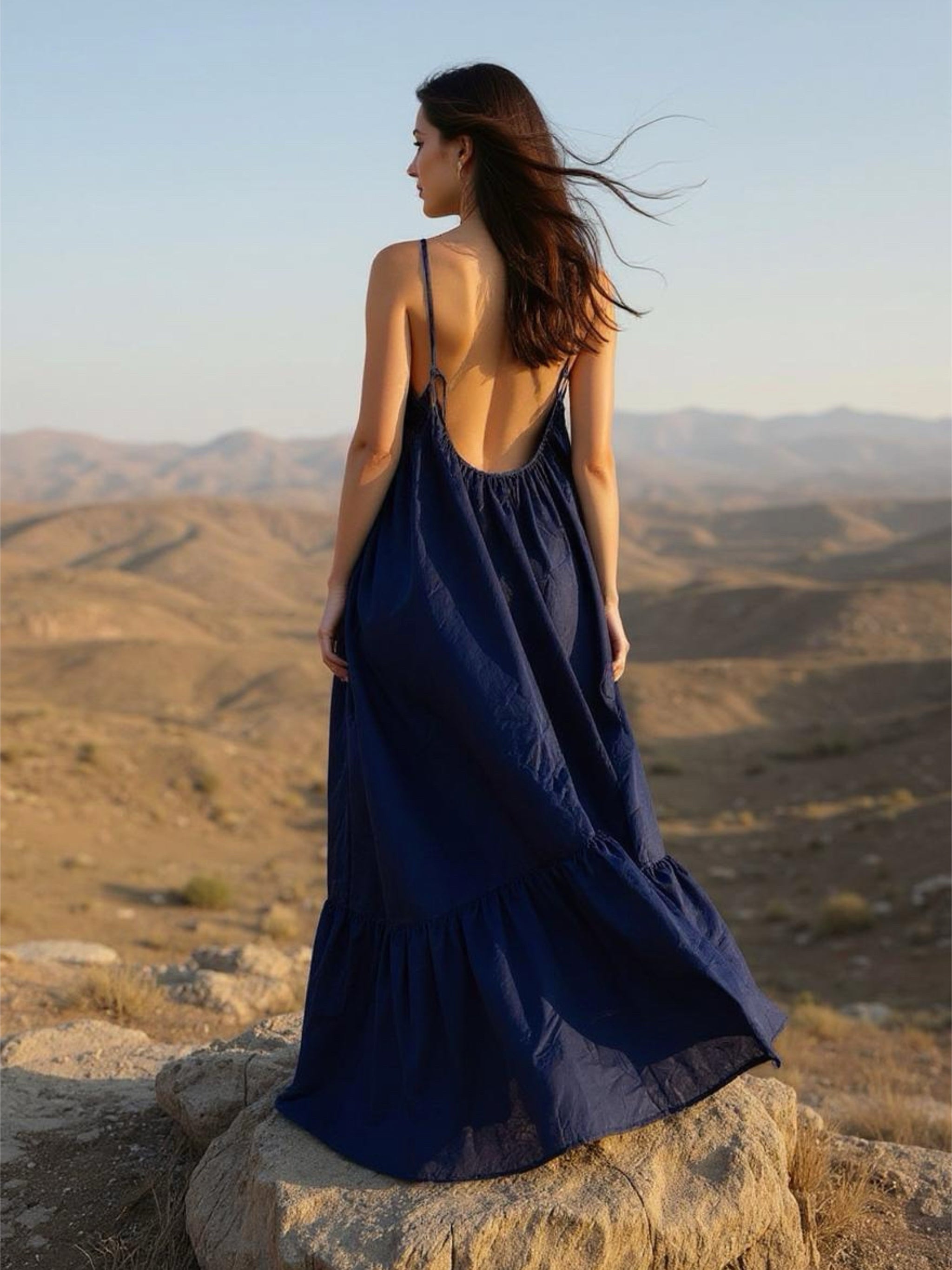 Woman wearing a navy backless maxi dress with adjustable tie-back straps, standing on a rocky hill with mountains in the background.