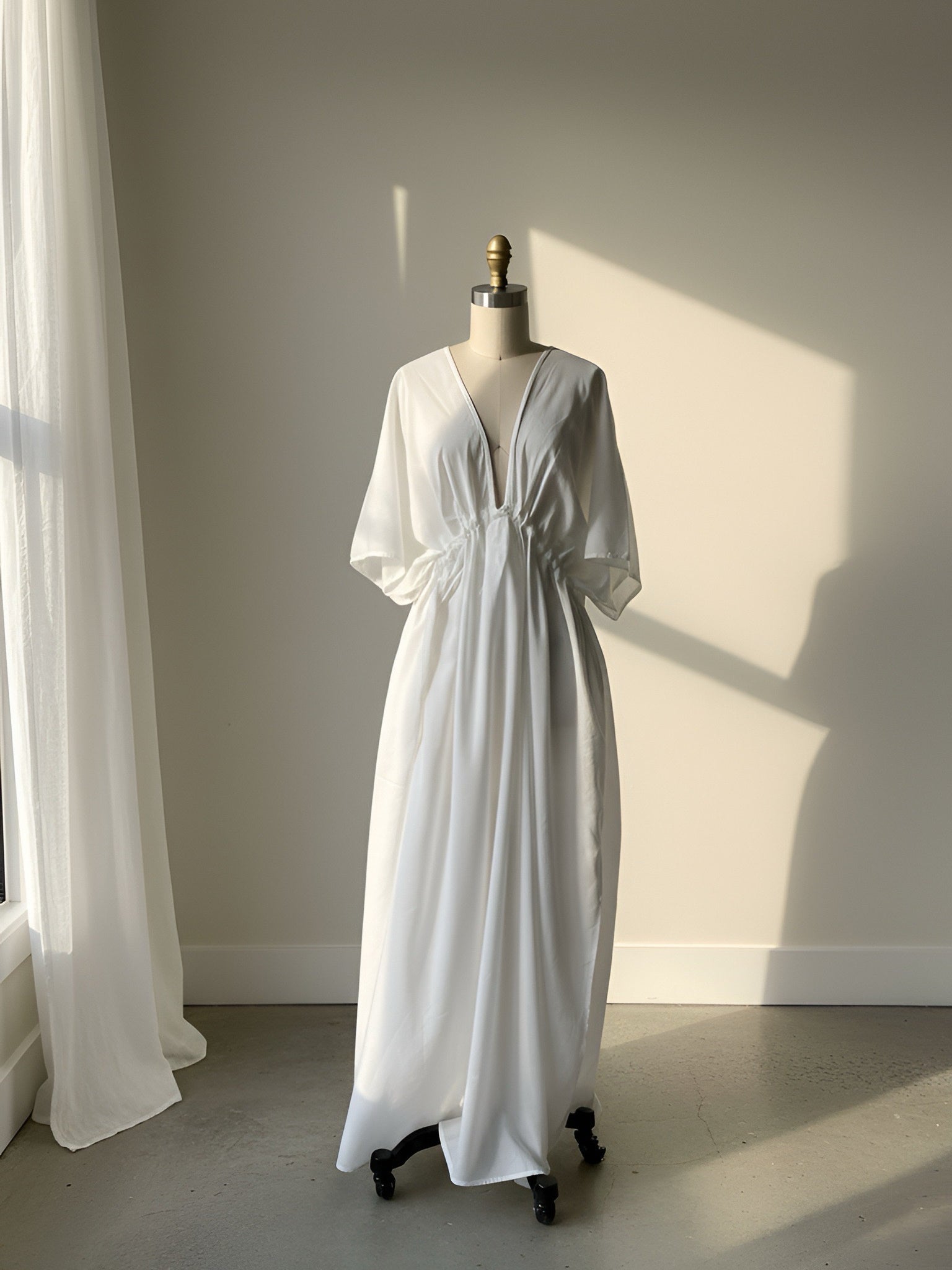 White kaftan dress on a mannequin against a neutral wall with sunlight casting shadows, resort dress for beach day.