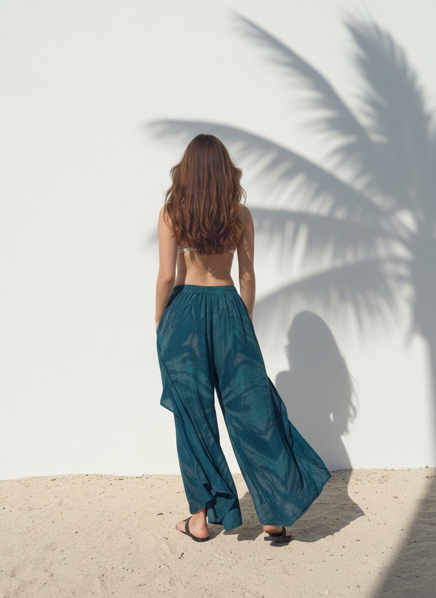 Woman wearing a white top and teal tie dye boho pants standing against a white wall with sand at the bottom, perfect beach outfit.