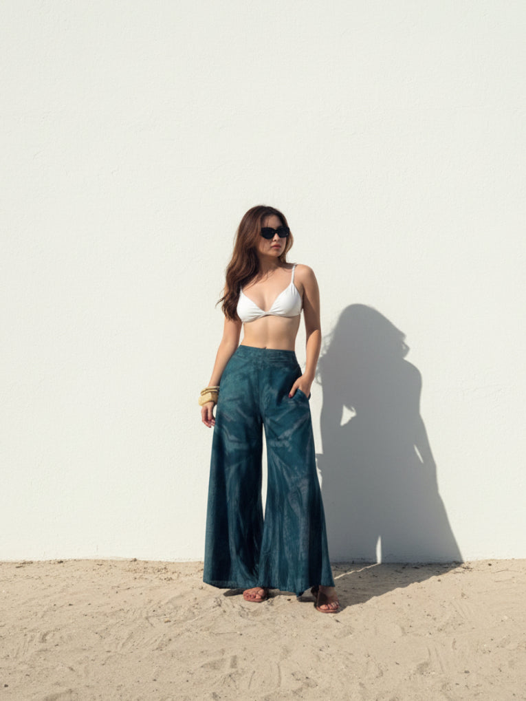 Woman wearing a white top and teal tie dye boho pants standing against a white wall with sand at the bottom, perfect beach outfit.