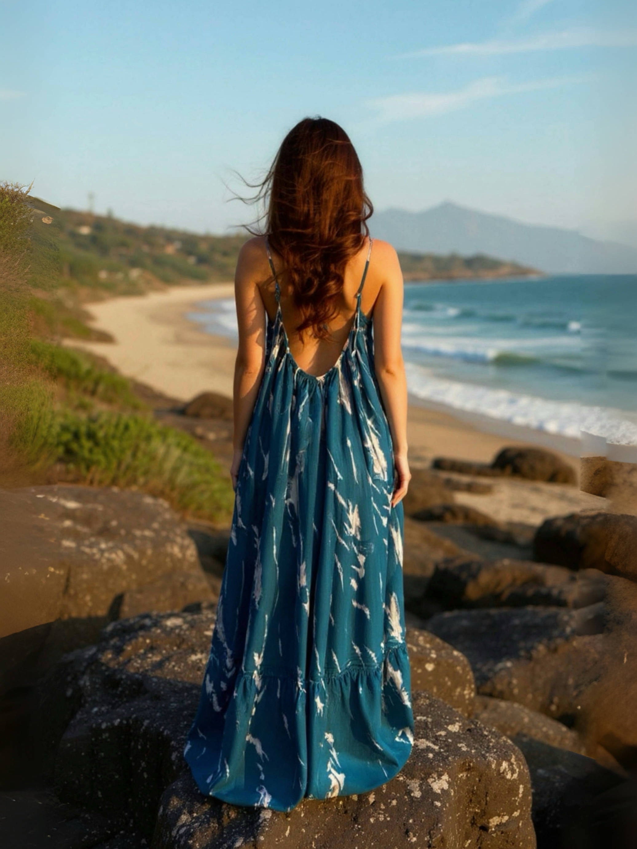 Woman in a teal backless maxi dress standing on a rocky beach with ocean and mountains in the background