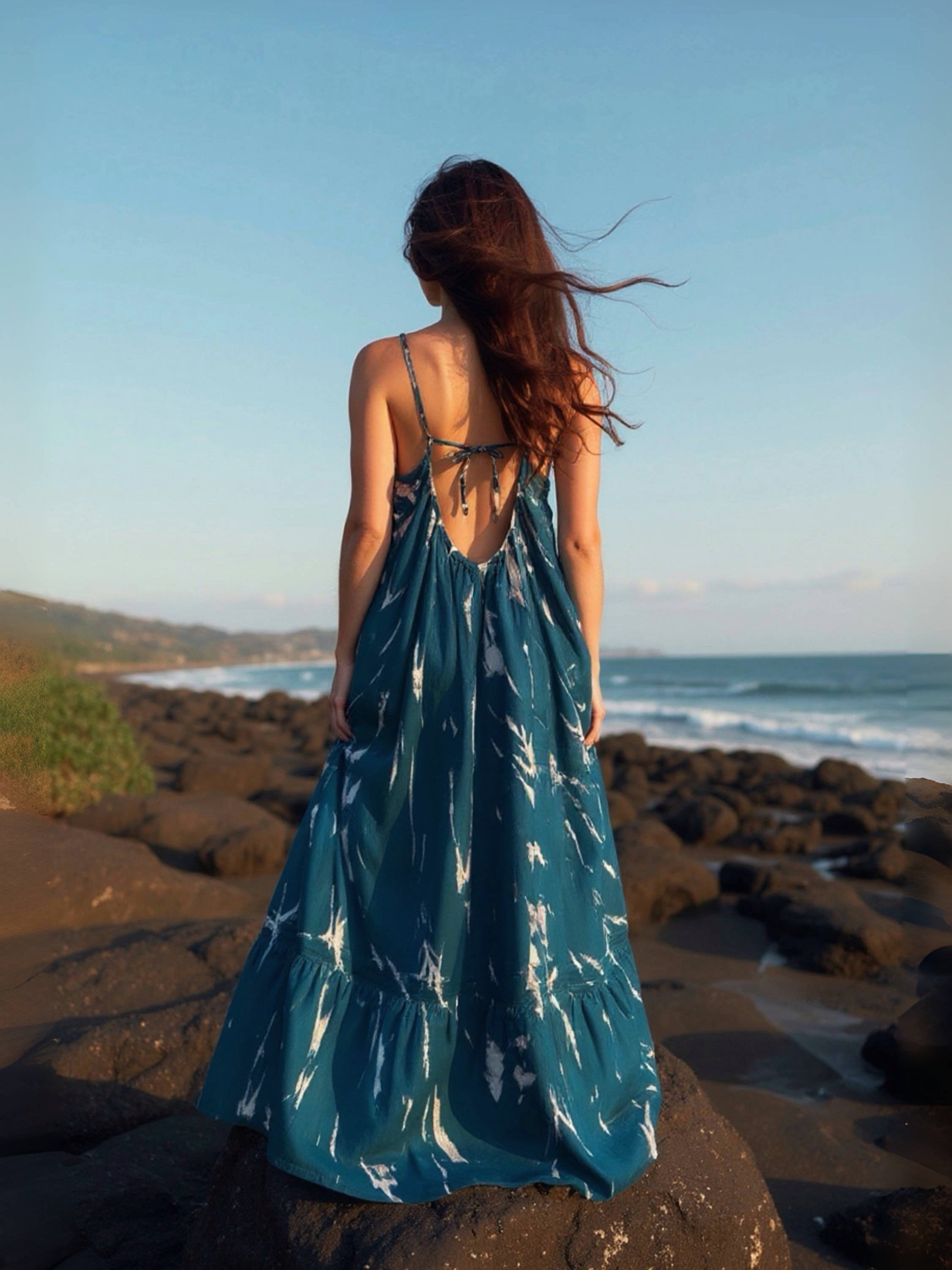 Woman in a teal backless maxi dress standing on a rocky beach with ocean and mountains in the background